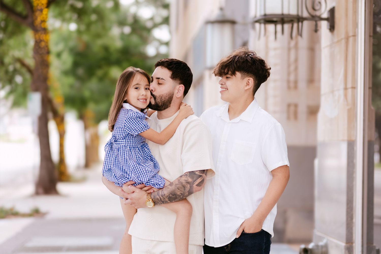 A father holds his young daughter in a blue dress while standing with his teenage son on a sunny urban sidewalk.
