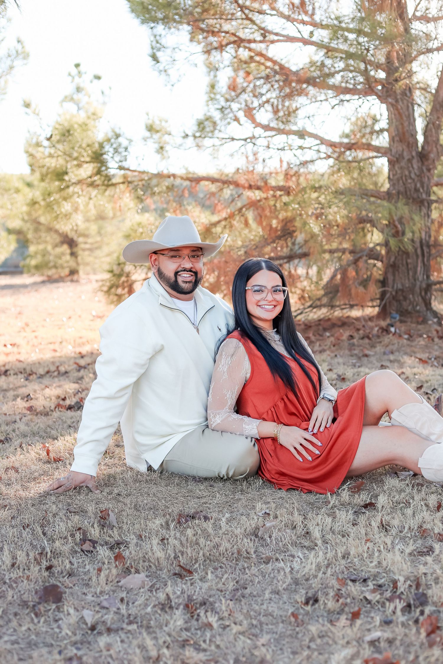 Smiling couple sitting on the ground outdoors against a pine tree, man in cowboy hat and woman in red dress.