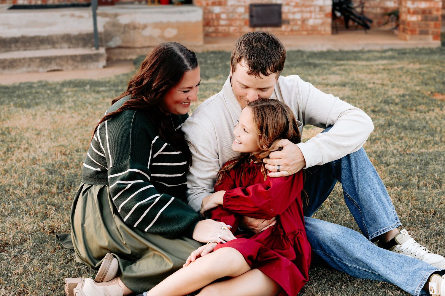 A family of three sits on grass laughing together, with a girl in a red dress between her parents.