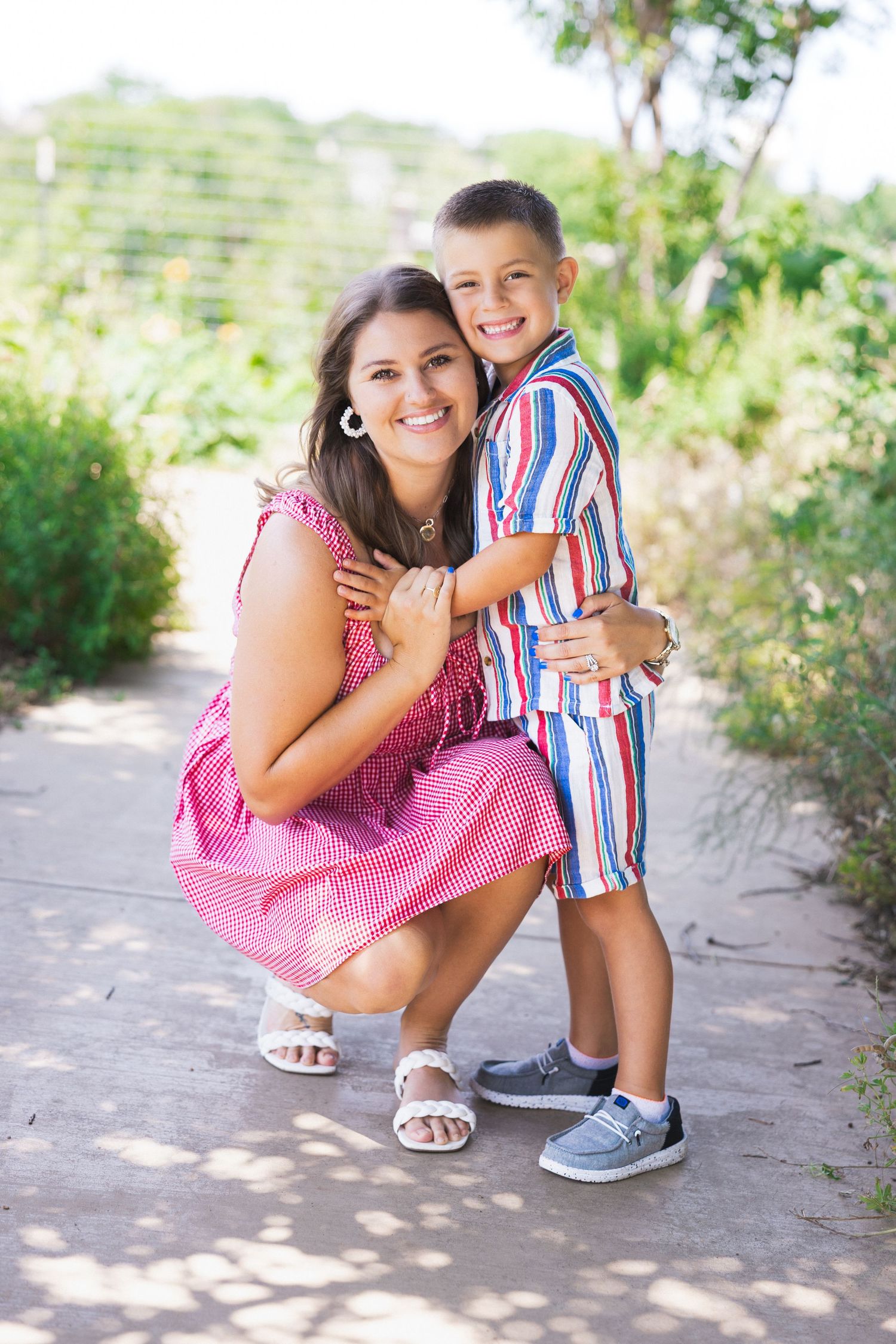A smiling mother crouches to hug her young son on an outdoor path.