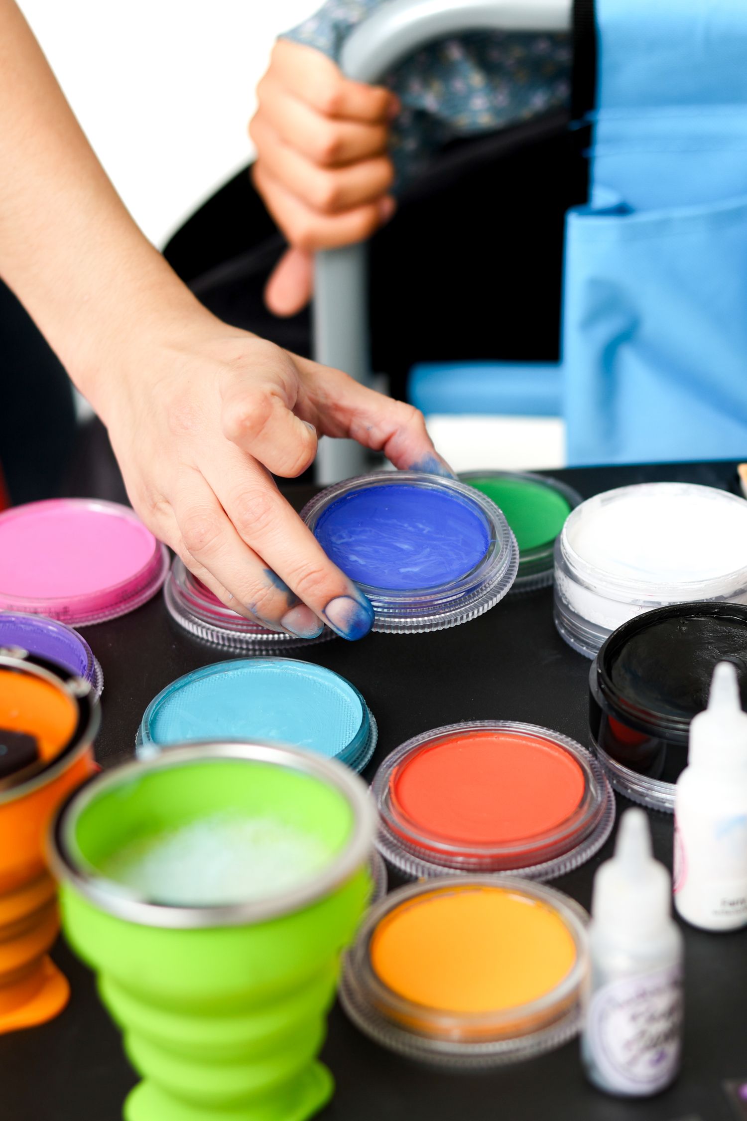Colorful face paint palette with blue, pink, green, and orange pots on a black table with a hand selecting a color.