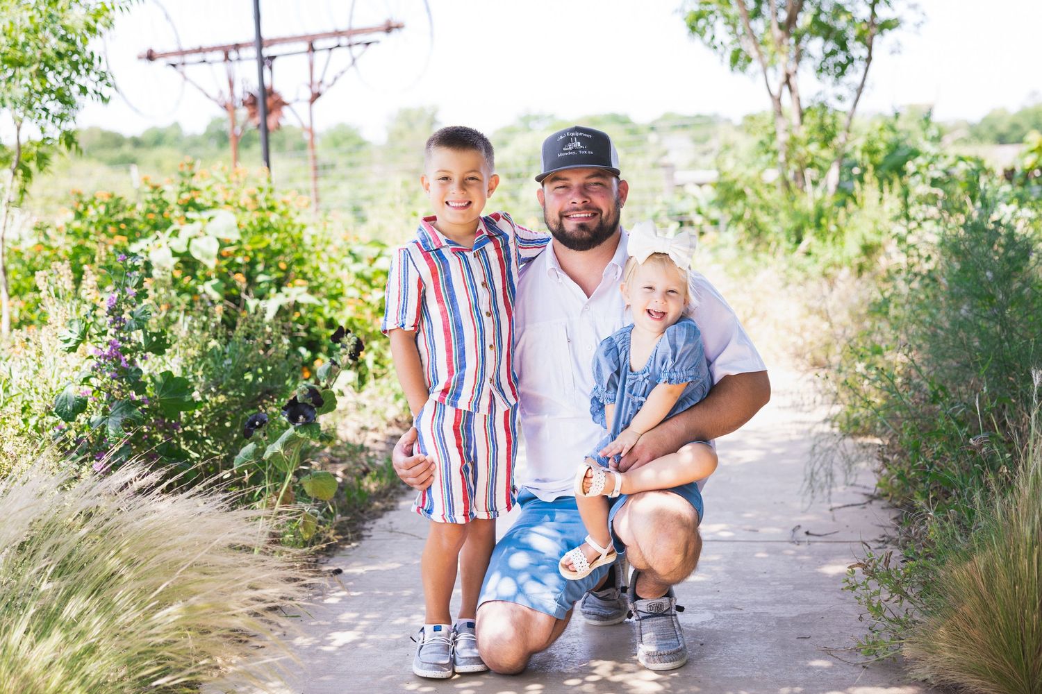 A father kneels on a garden path with his two young children, smiling for a family portrait.