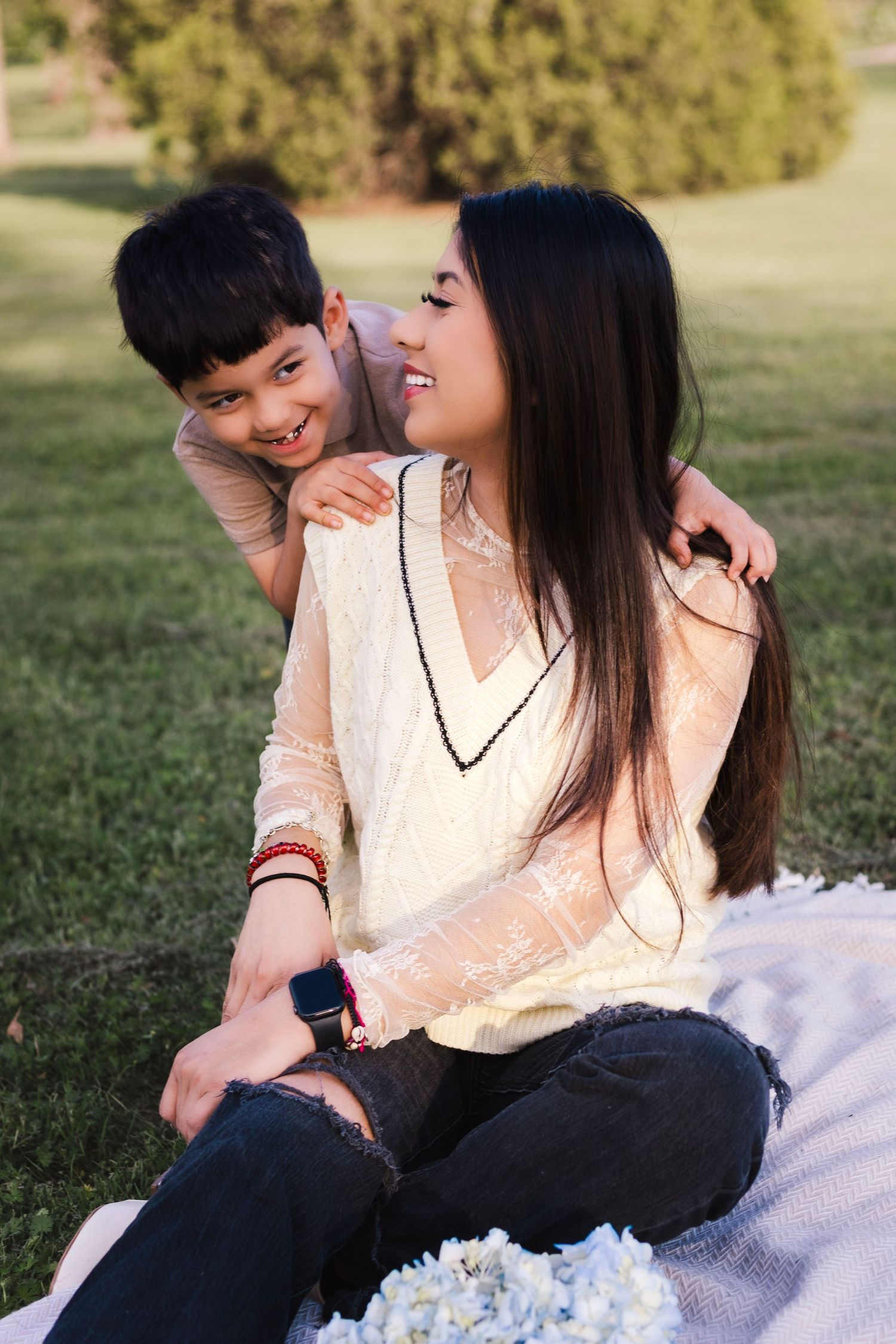 A young Asian woman sits on a blanket in a park while a smiling boy hugs her from behind on a sunny day.