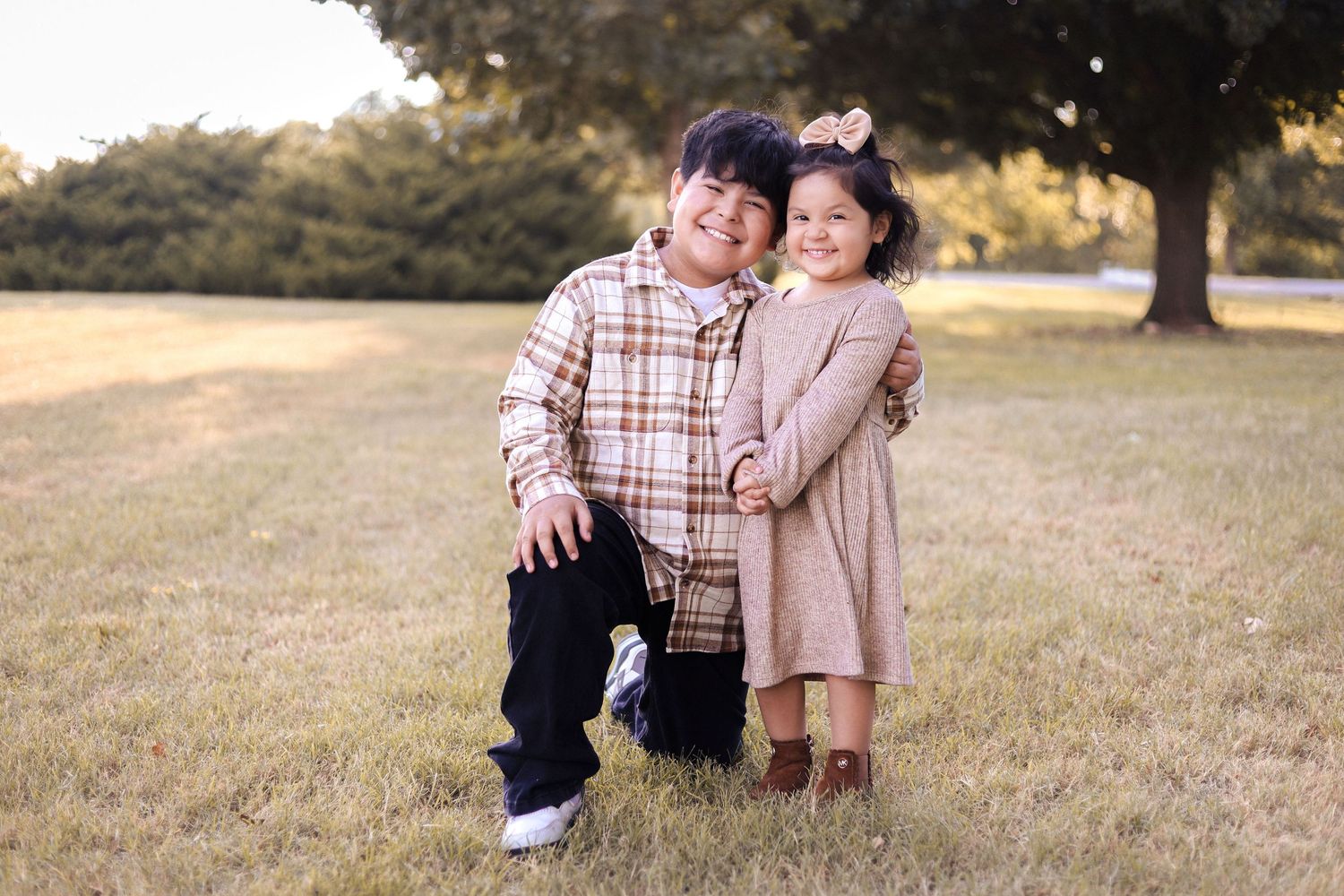 A boy and his little sister pose together affectionately in a grassy park for a sibling portrait.