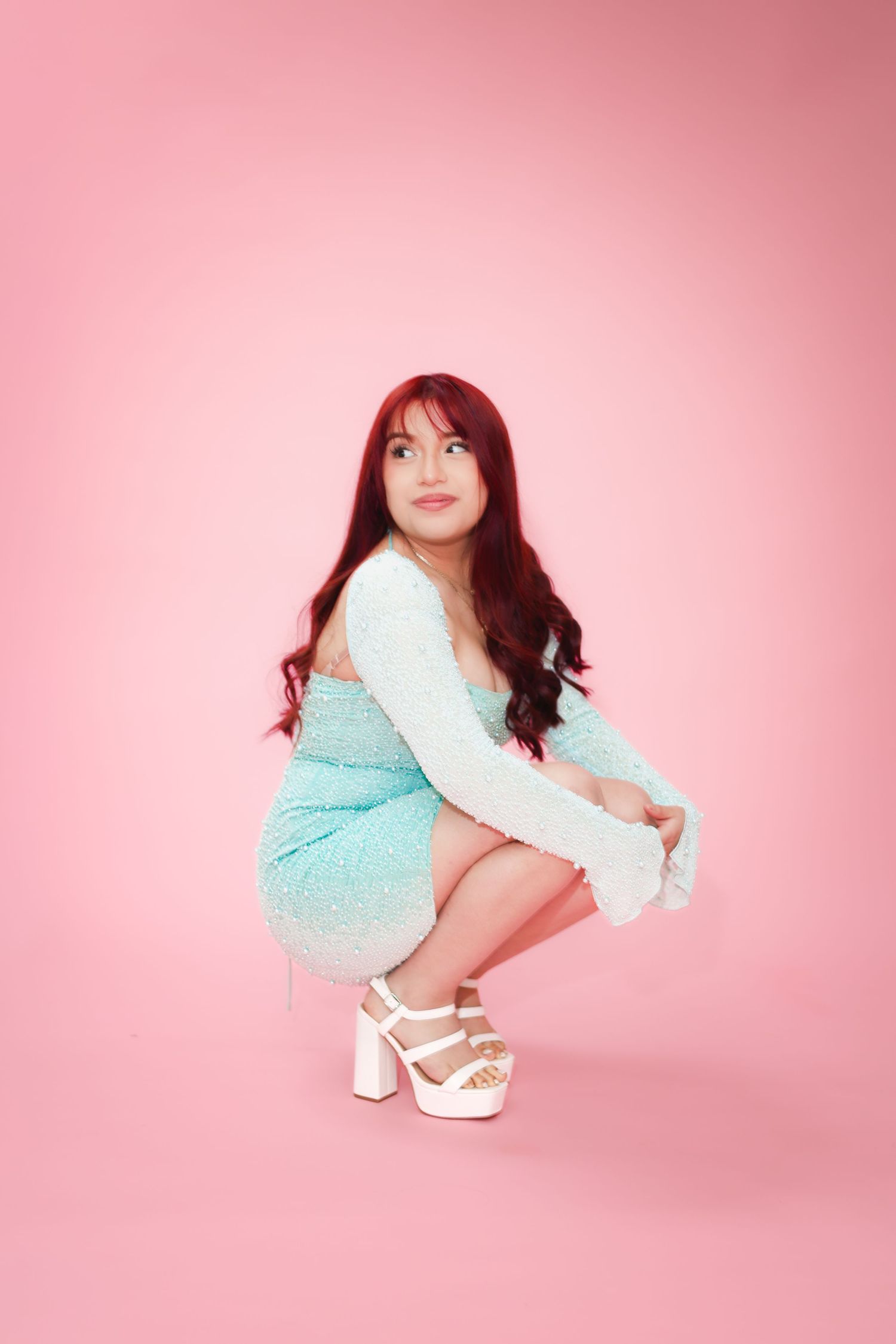 Young woman with red hair in a blue sequin dress crouching in white platform heels on pink background.
