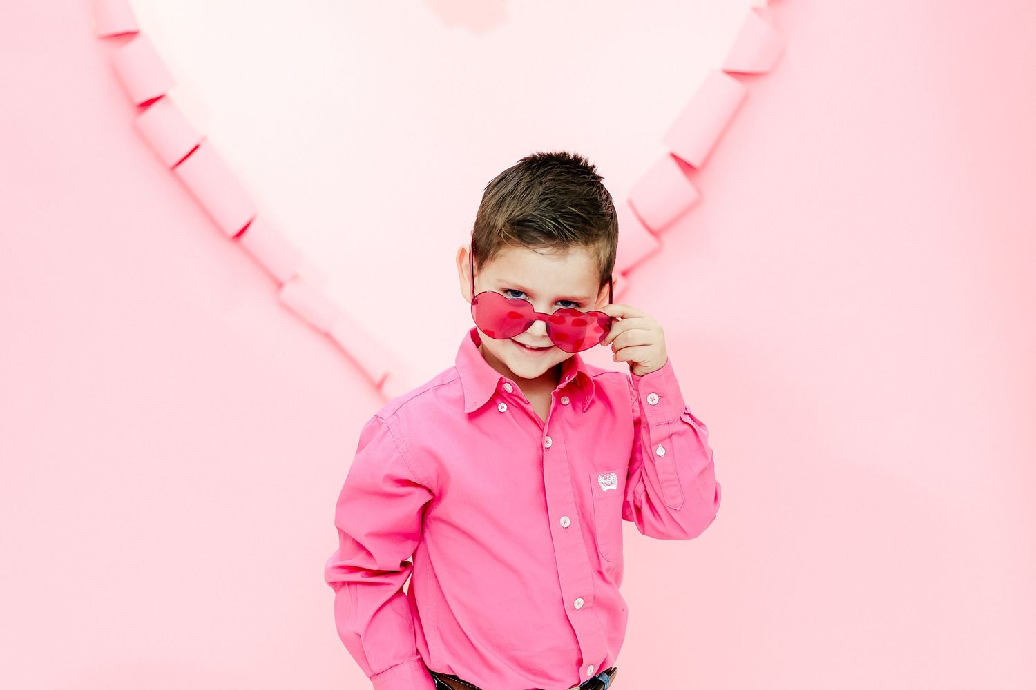 Cute boy in pink shirt and heart-shaped sunglasses posing in front of a pink heart background.