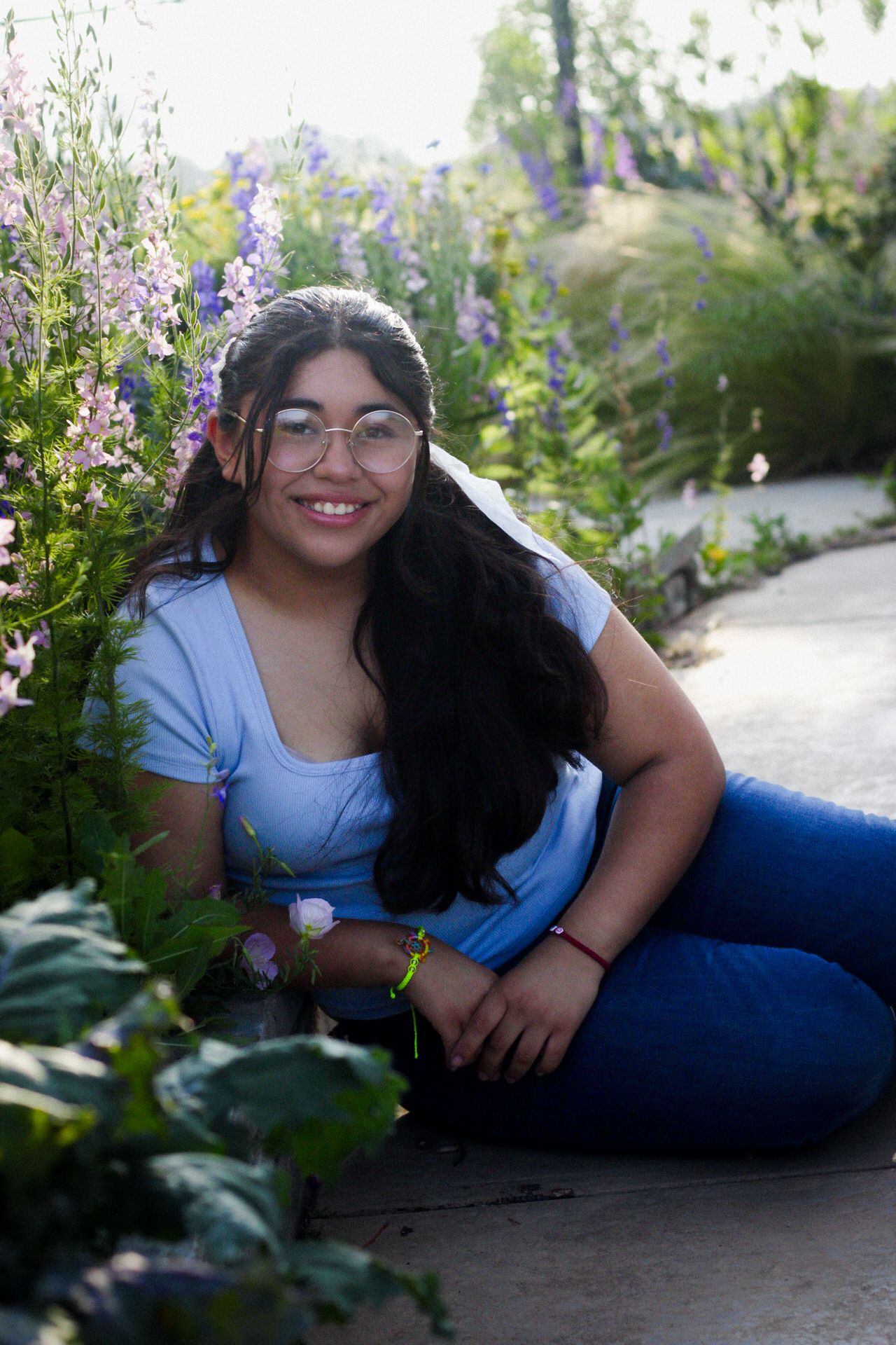 A teenage girl with long dark hair and glasses sits among colorful purple flowers in a garden.