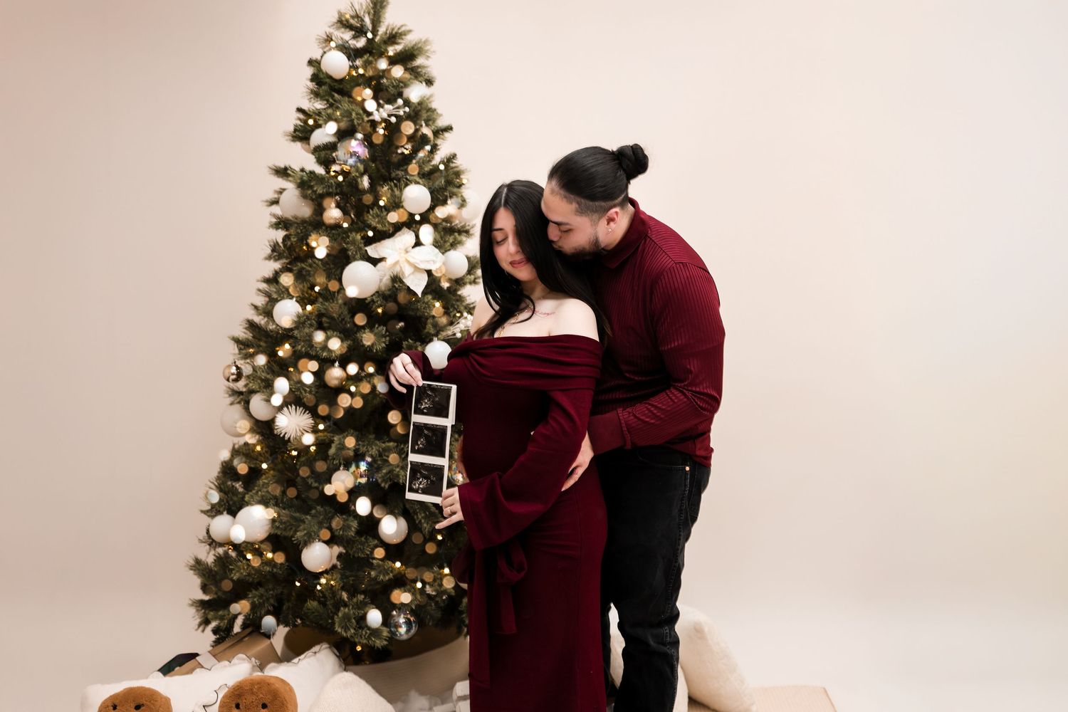 Pregnant woman in burgundy gown holds ultrasound photos while partner kisses her head by Christmas tree.