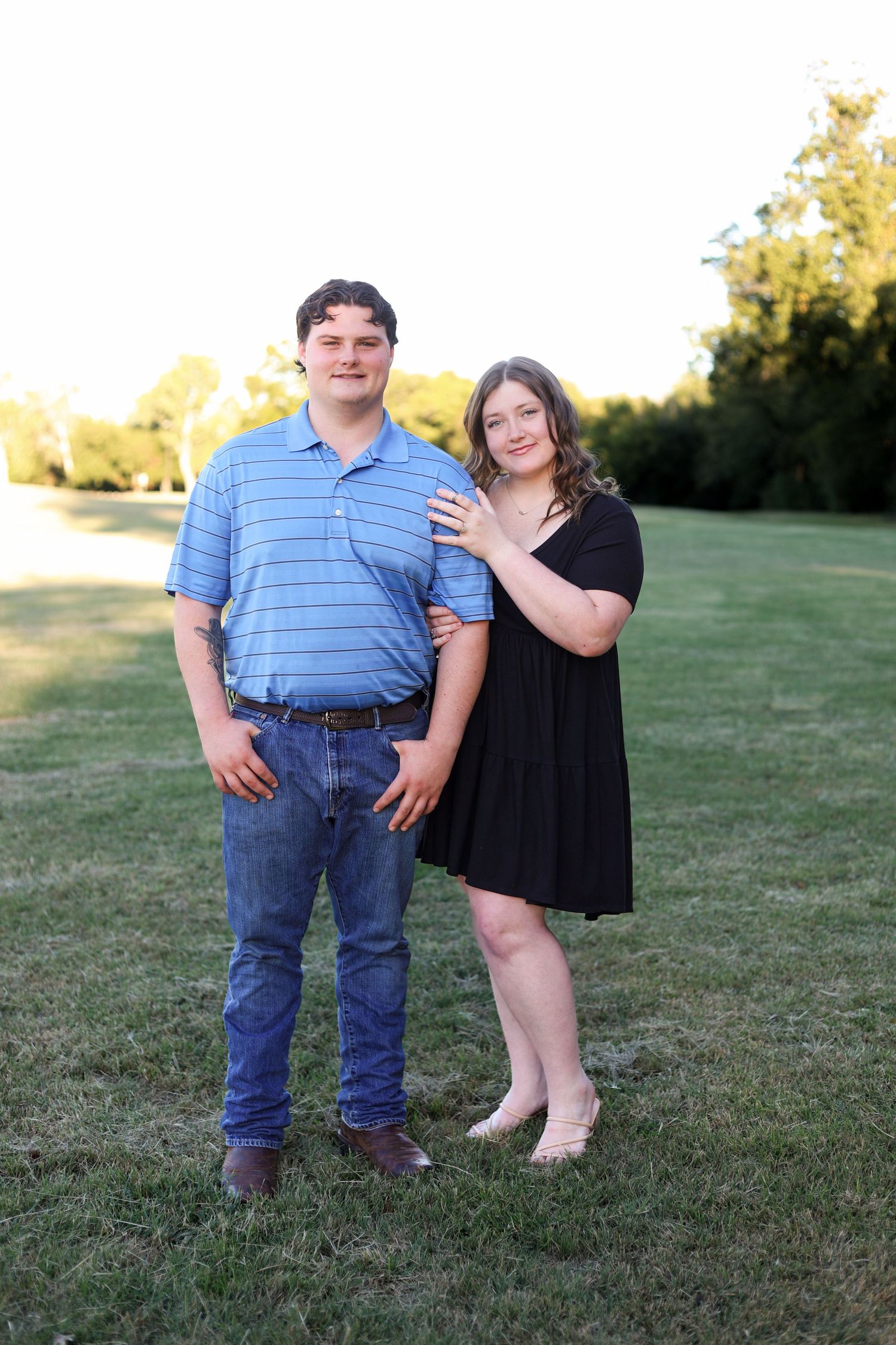 Young couple posing together on green lawn at sunset, man in blue polo and woman in black dress.