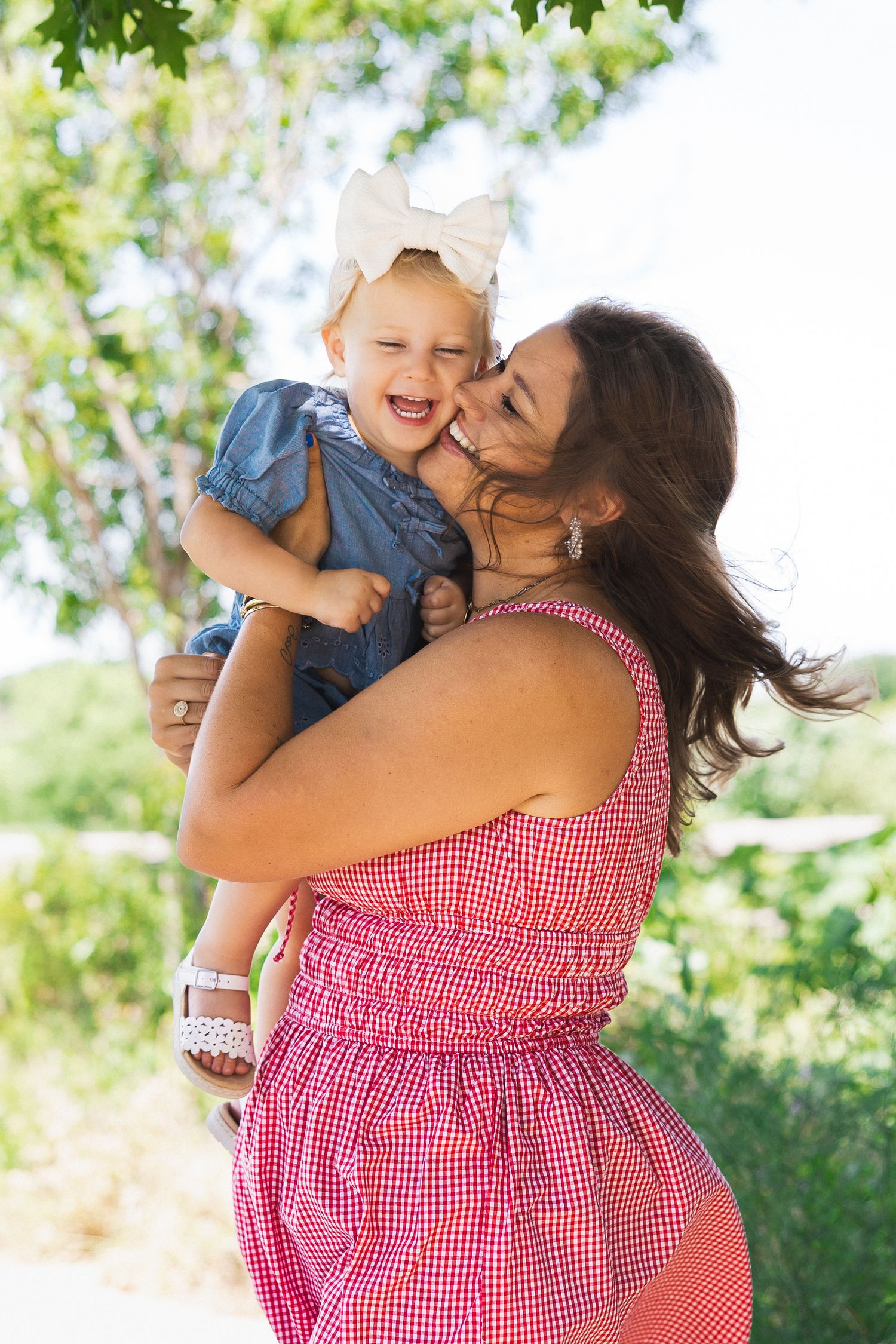 A mother in a pink dress kisses her laughing baby girl with a white bow outdoors.