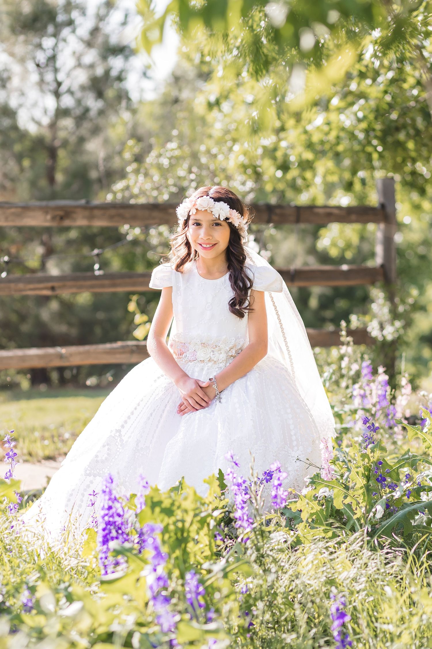 Young girl in white First Communion dress and floral crown poses among purple wildflowers in a sunny garden.