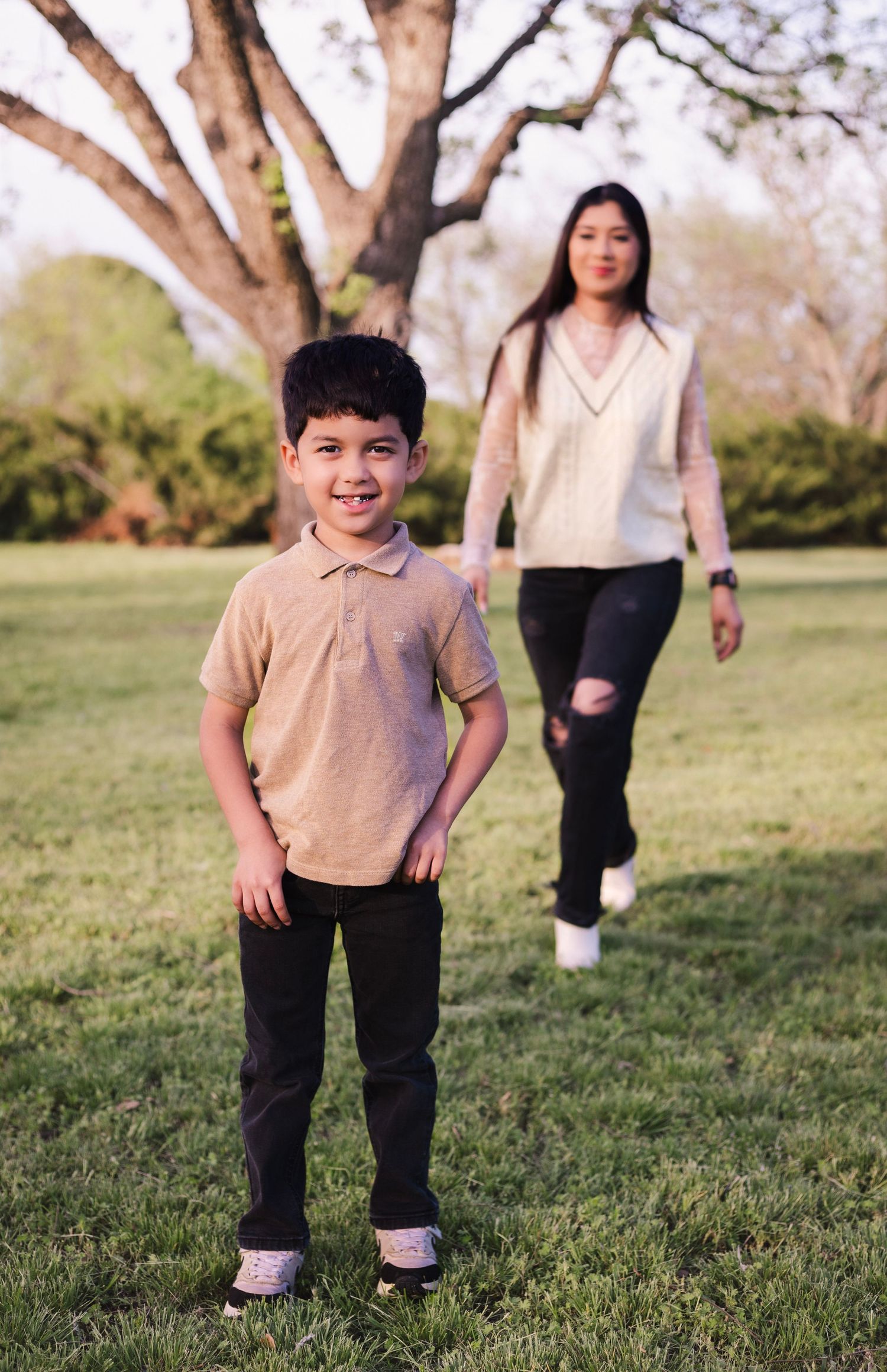 A cheerful boy poses in a park while his mother walks toward him in the background.