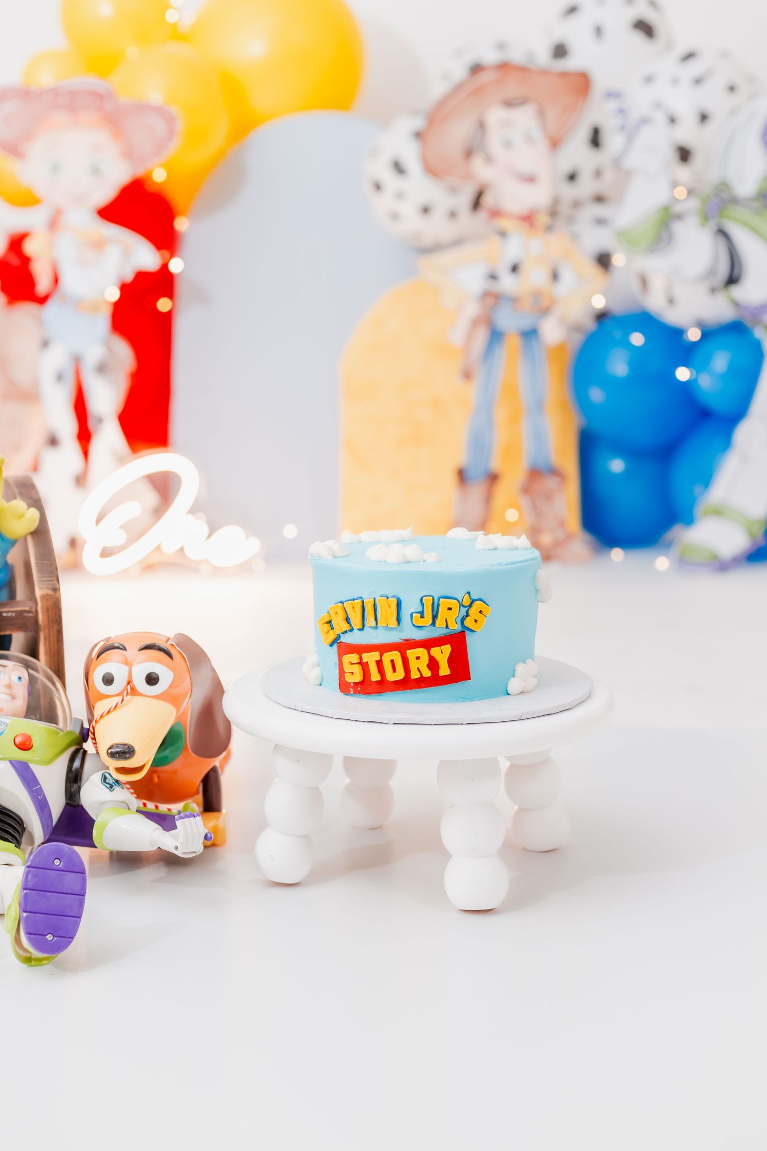 Toy Story themed first birthday cake on white stand with Slinky Dog figurine and Buzz Lightyear toy decorations.