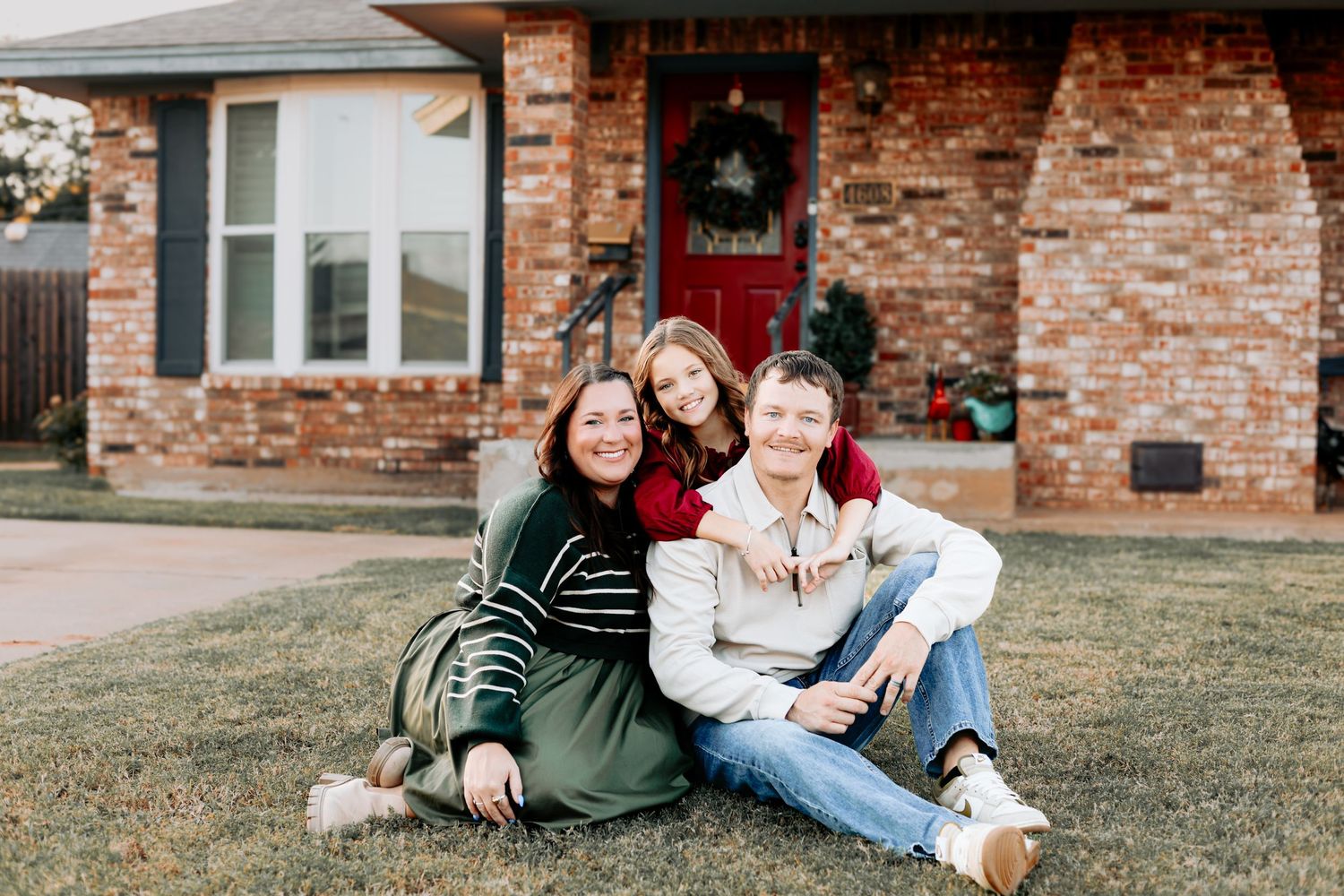 A happy family of three sits on a lawn in front of a brick house during outdoor photo session.