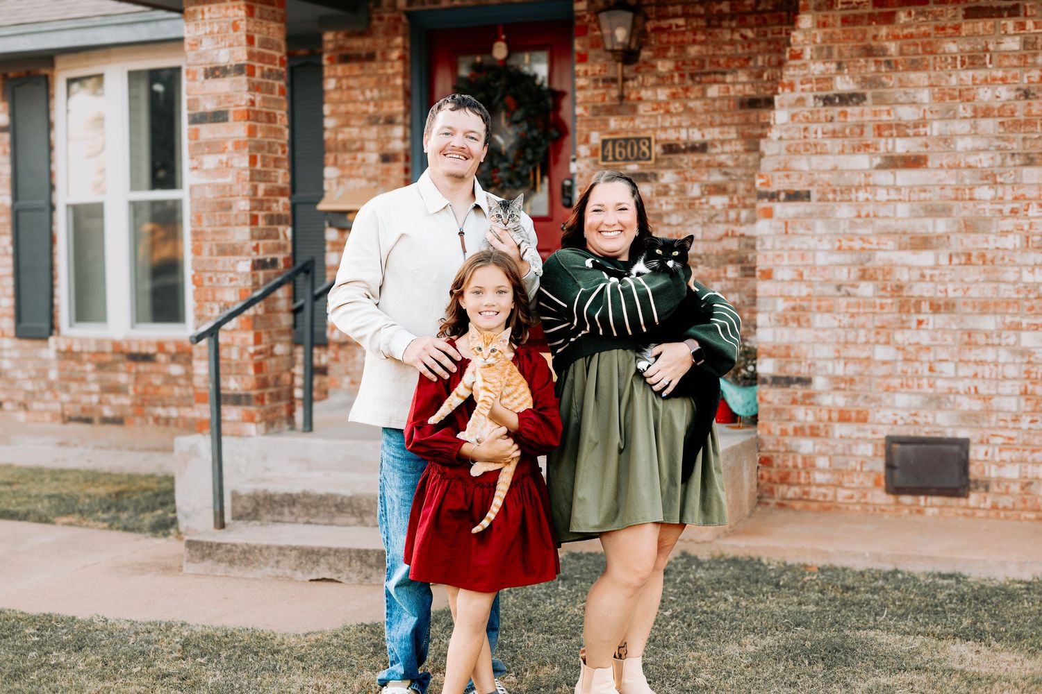 A family smiles outdoors holding cats in front of a brick house decorated for the holidays.