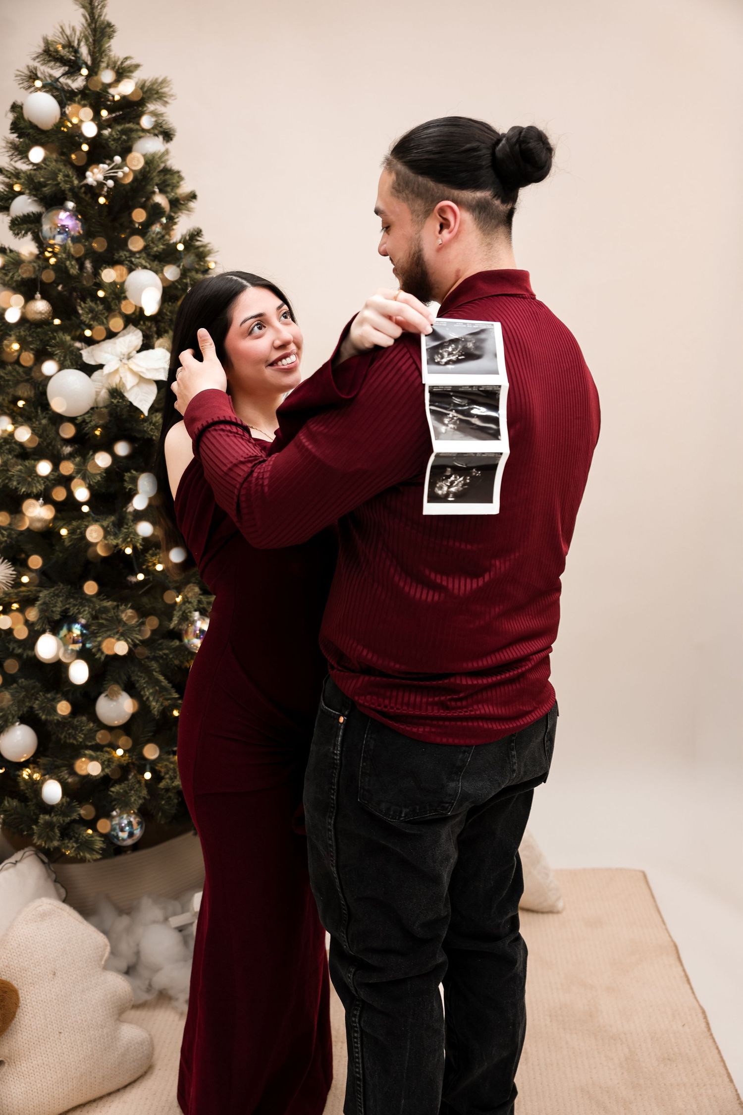 Couple embraces near Christmas tree as woman holds ultrasound photos behind mans back.