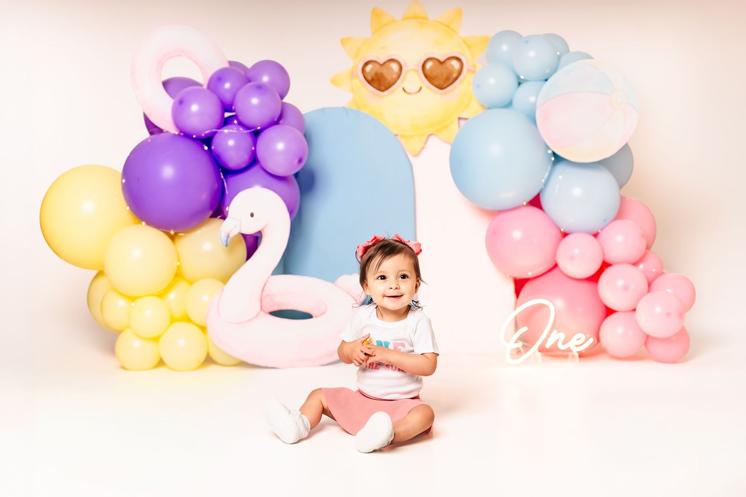 Baby girl sits among colorful pastel balloons and swan float for a whimsical first birthday cake smash photo session.