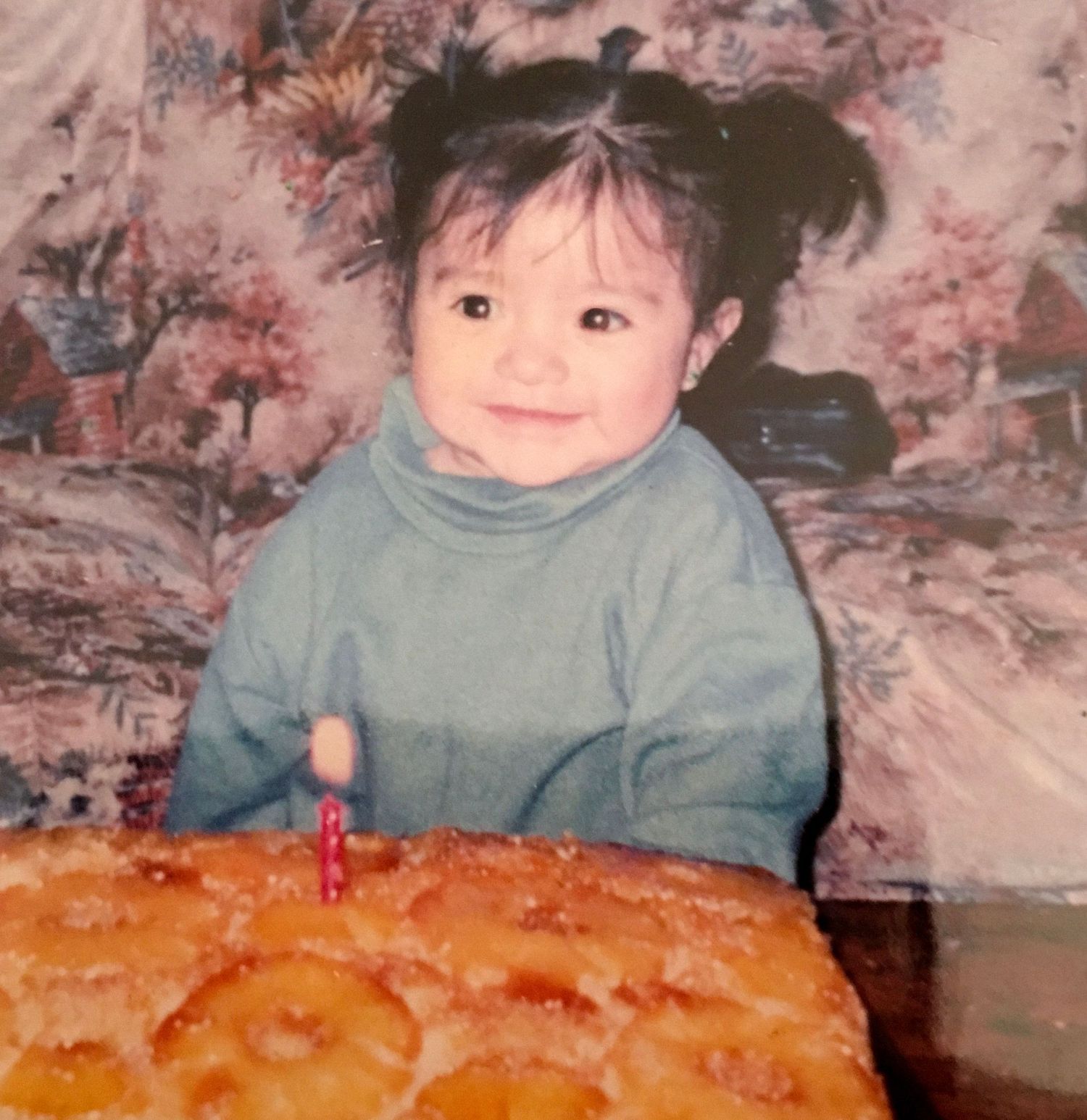 A smiling toddler in a blue turtleneck sits behind a pineapple upside-down cake with a candle on their birthday.