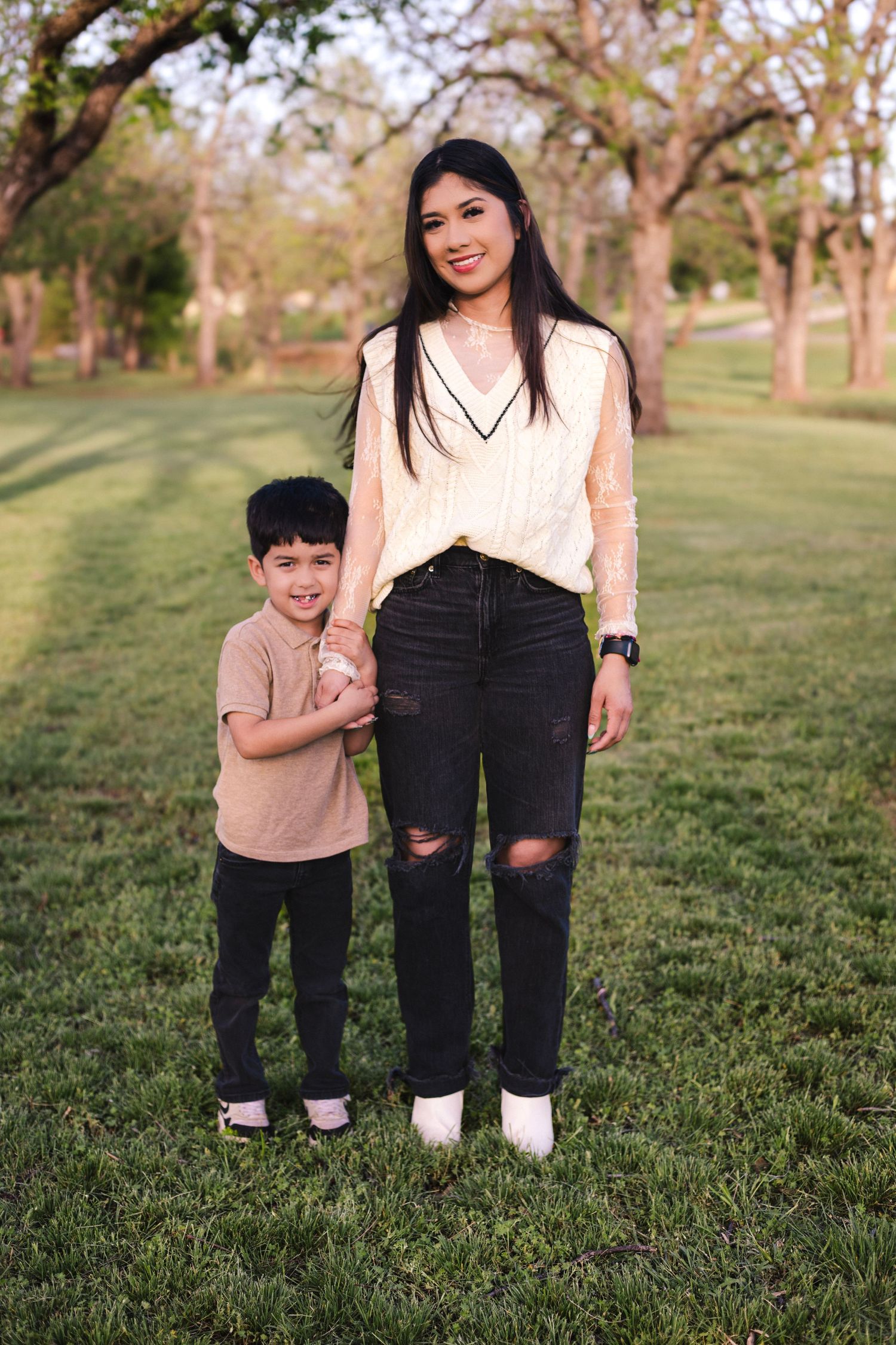 A smiling woman in black jeans and white top holds hands with a young boy in a sunny green park.