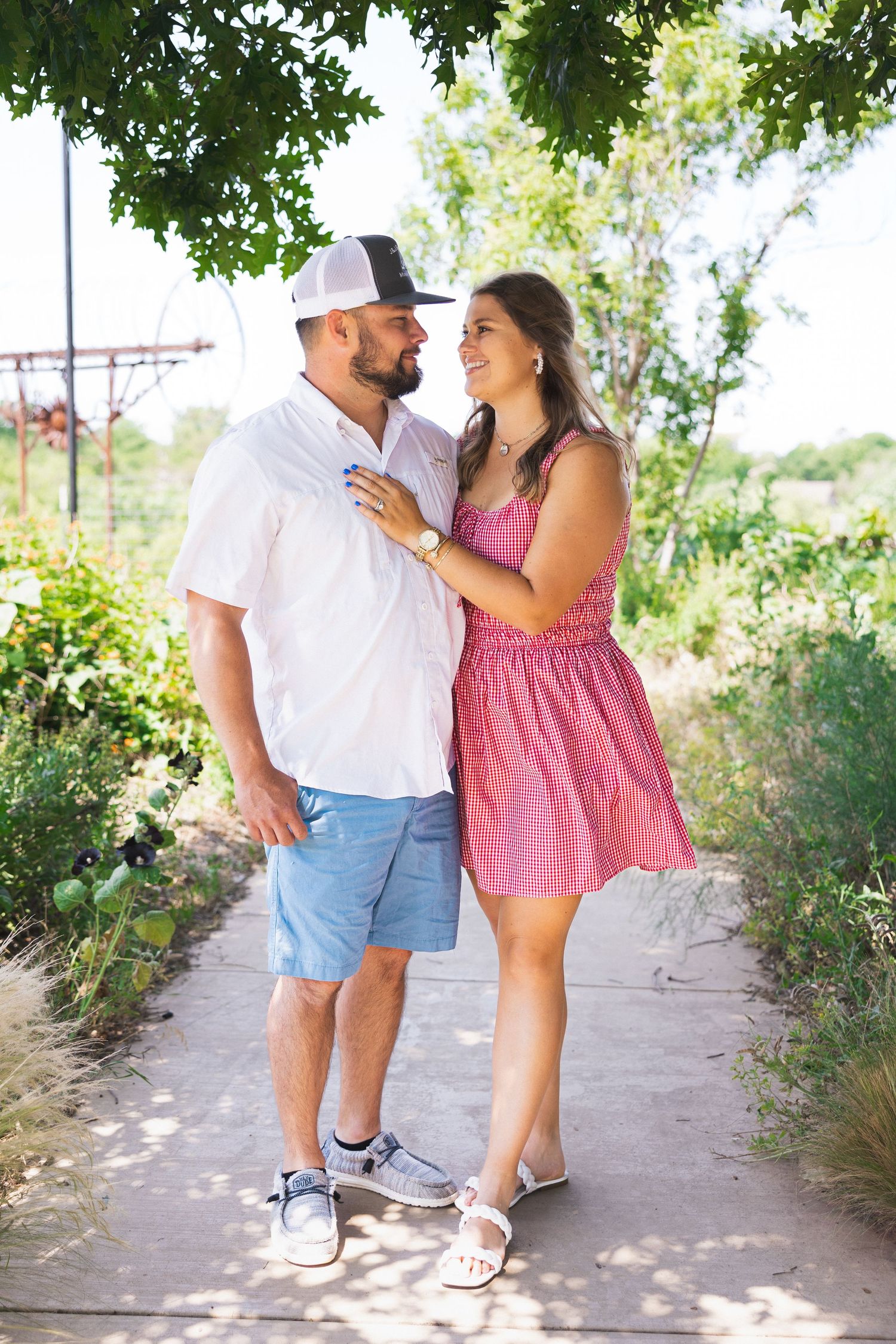 A couple in summer outfits stands close together smiling under green trees on a sunny path.