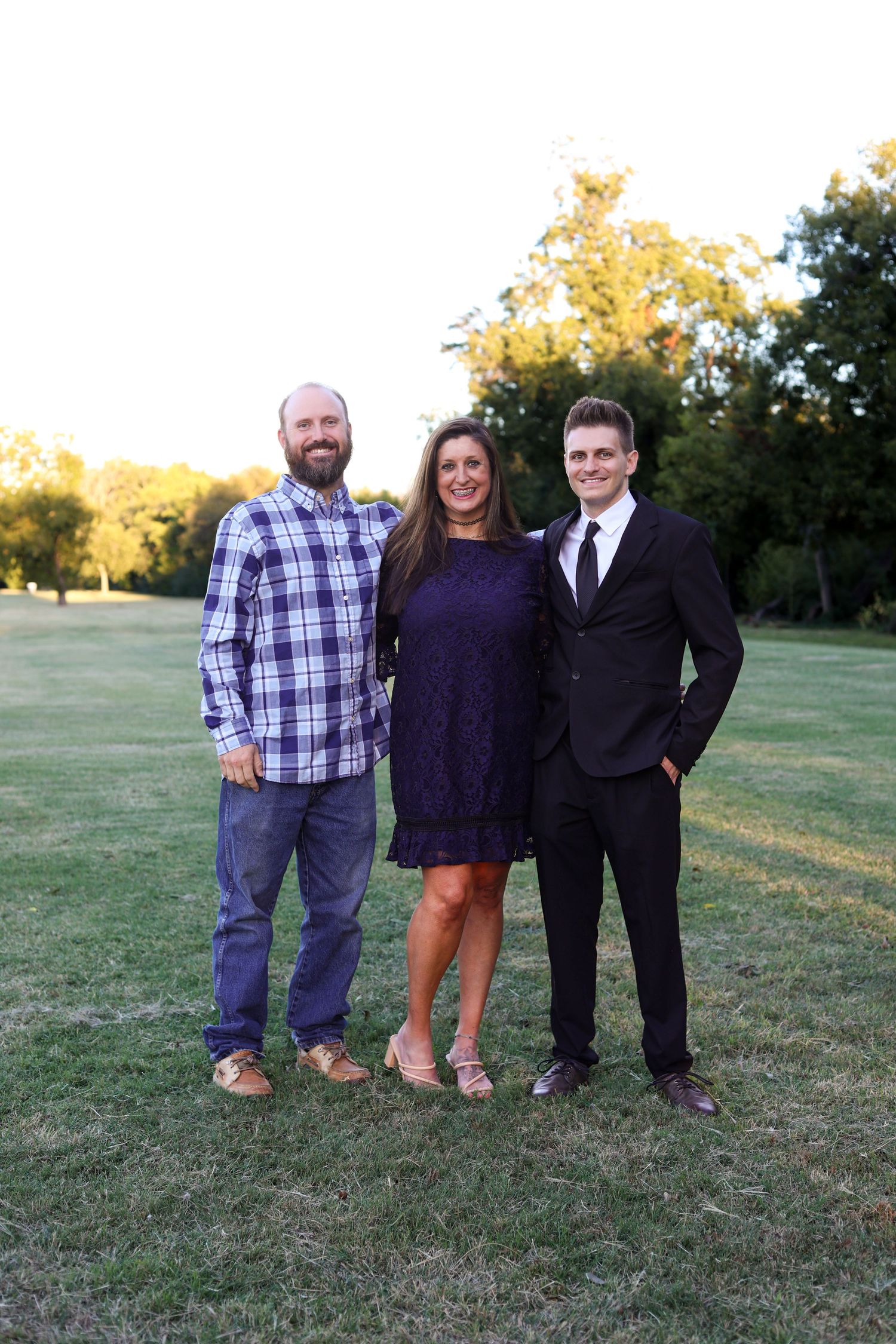 Three family members smiling together outdoors on a green lawn at golden hour.