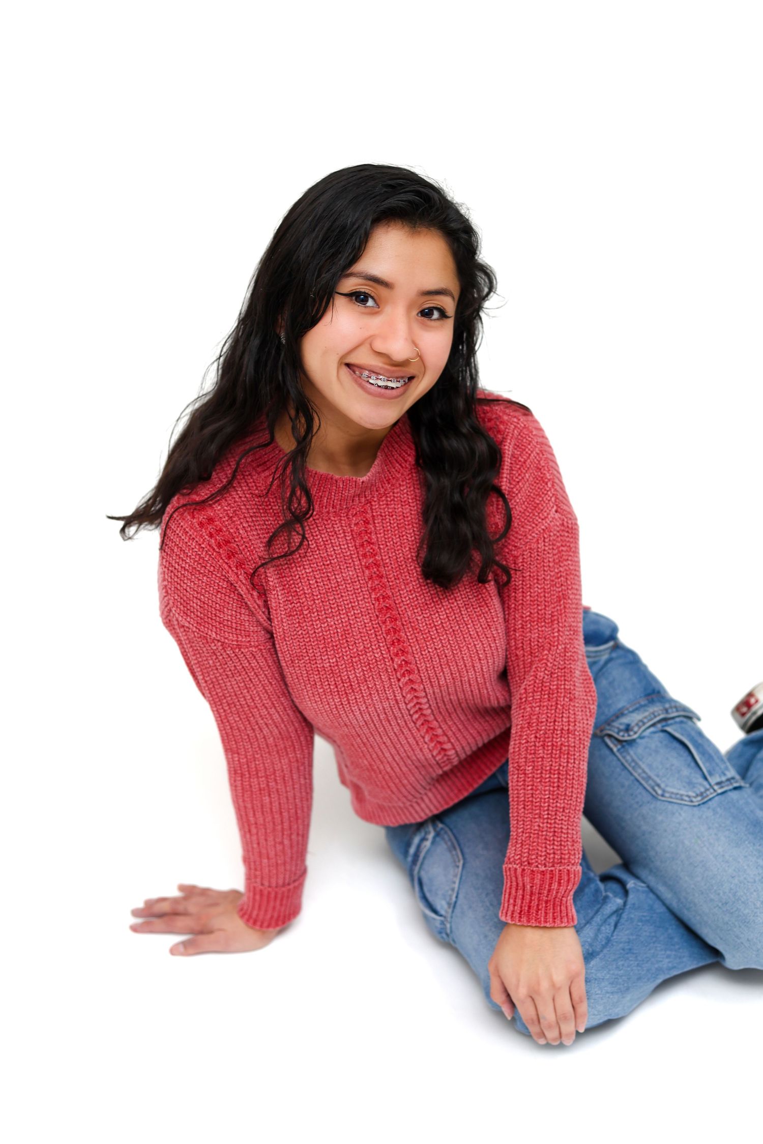 Smiling teenage girl with braces wearing a red sweater and jeans sitting on a white background.
