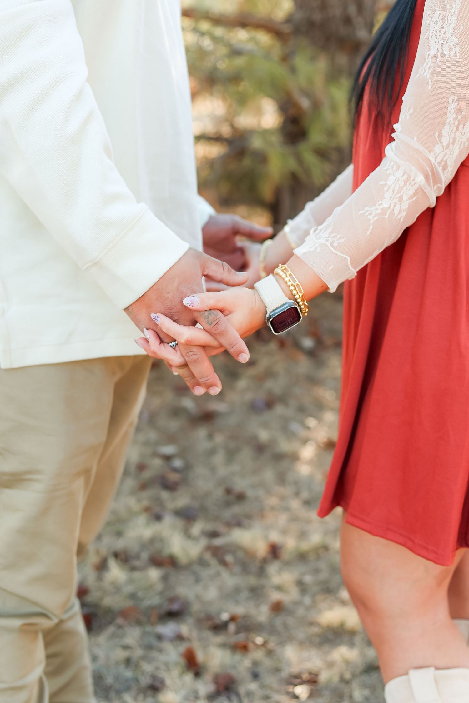 Close-up of couple holding hands outdoors, woman wearing gold bracelets and smart watch in red dress.