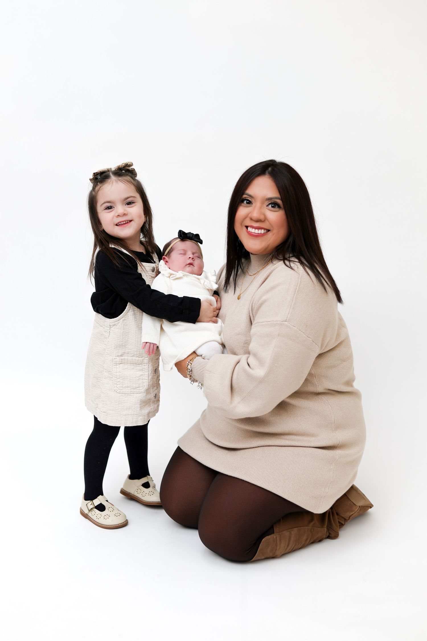 Smiling mother kneels holding newborn baby while young daughter stands beside them on white background.