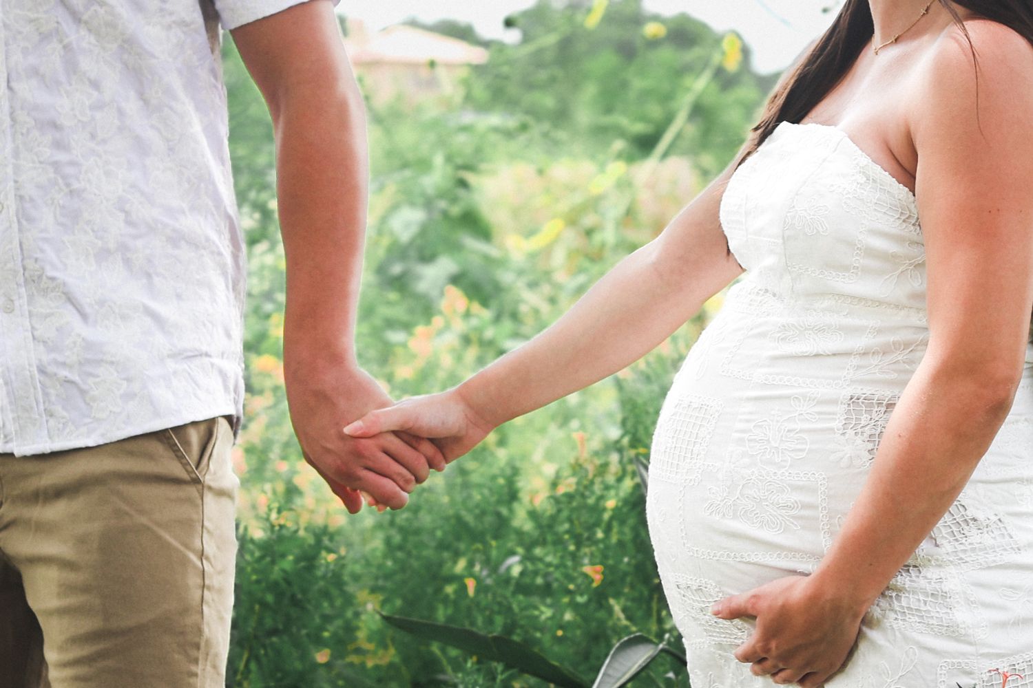 Pregnant woman in white dress holding hands with partner outdoors in green garden.