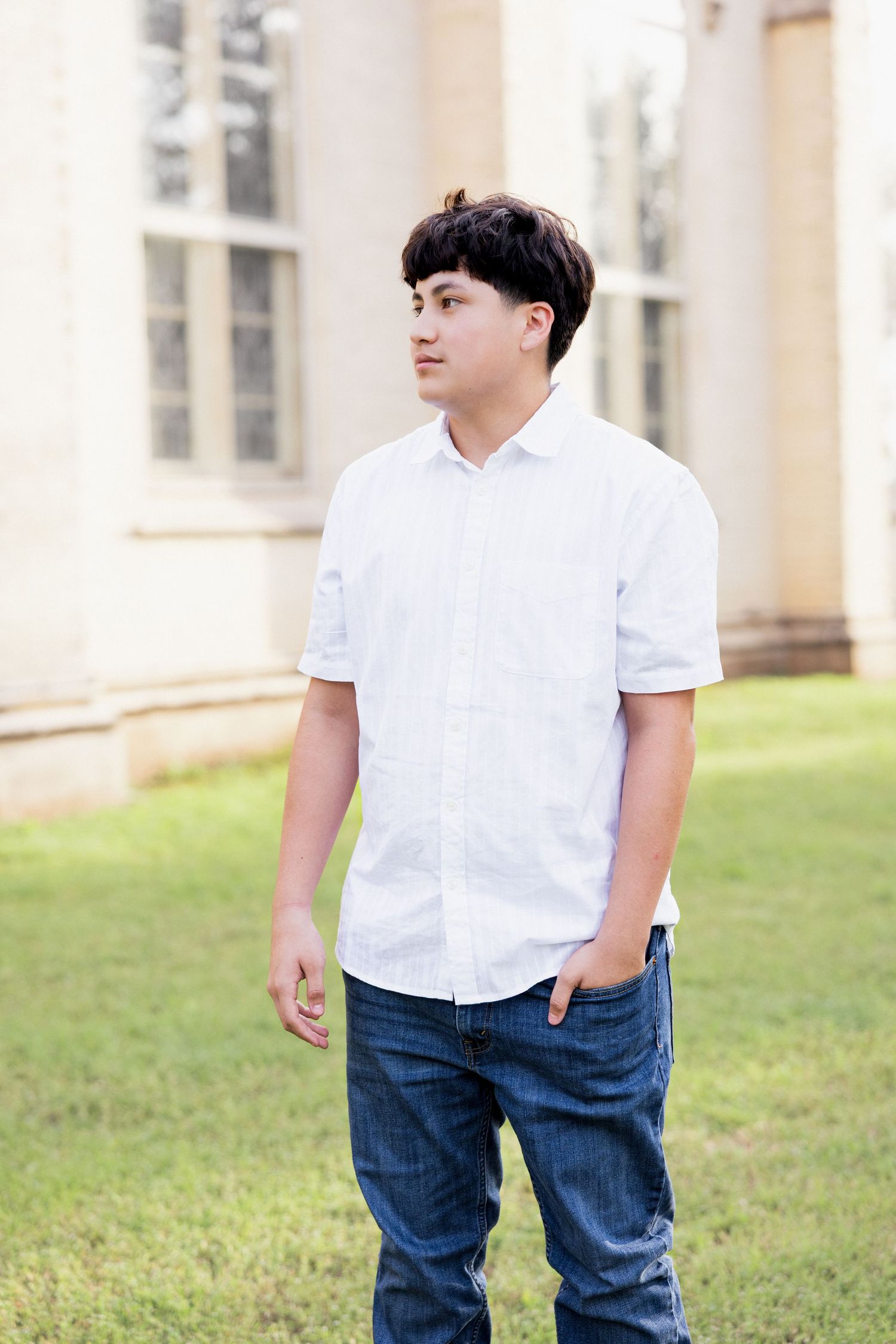 Teenage boy in white short-sleeve shirt and jeans stands outdoors near a classic stone building.