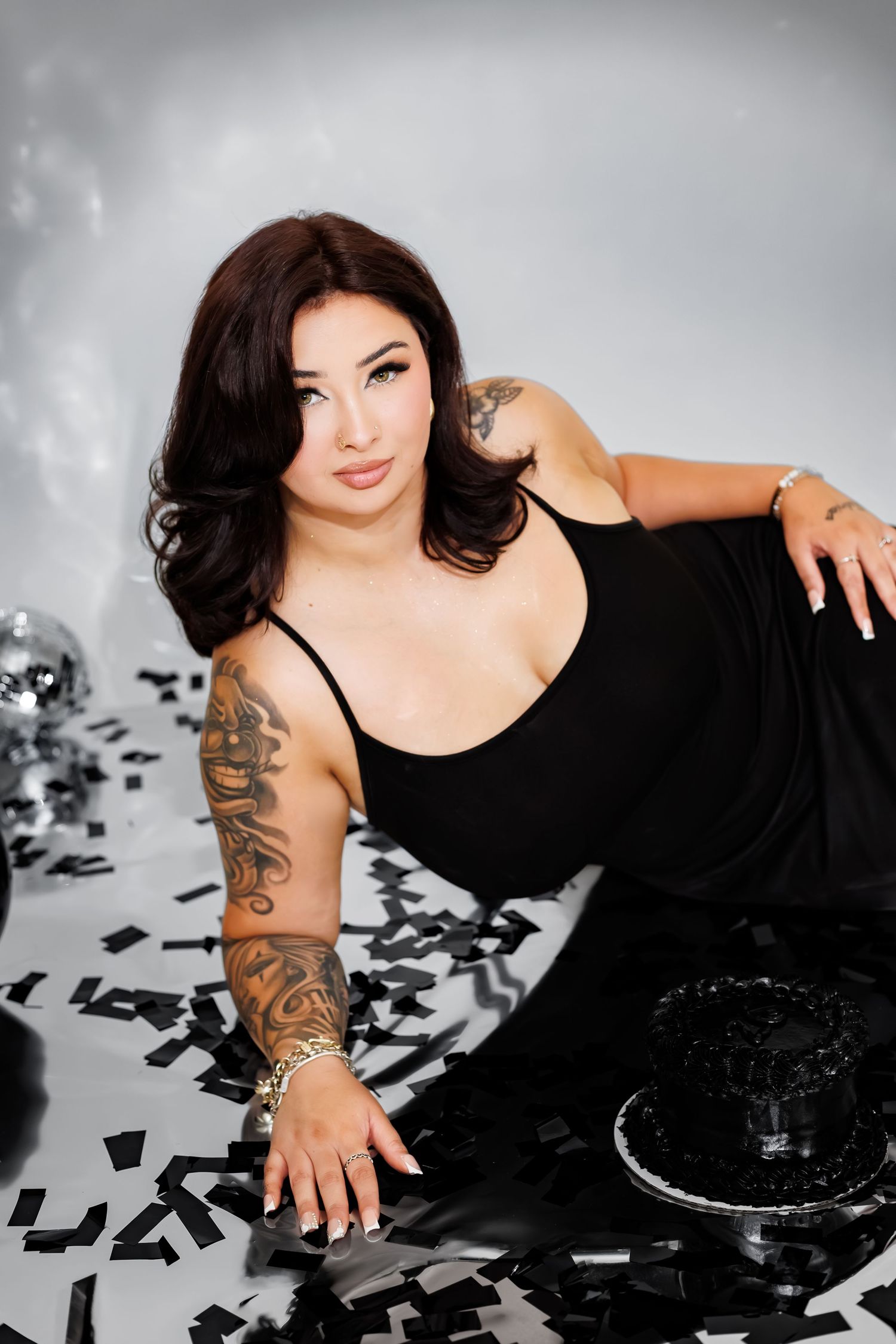 Woman in black dress with tattoos poses on floor with confetti and disco balls in a studio setting.
