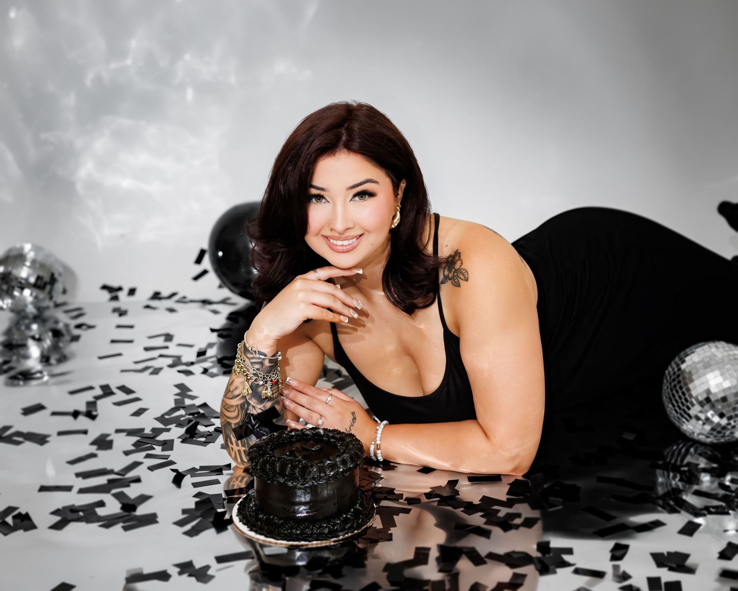 Woman in black dress lying on floor with black cake, confetti, and disco balls in a glamorous birthday photo shoot.