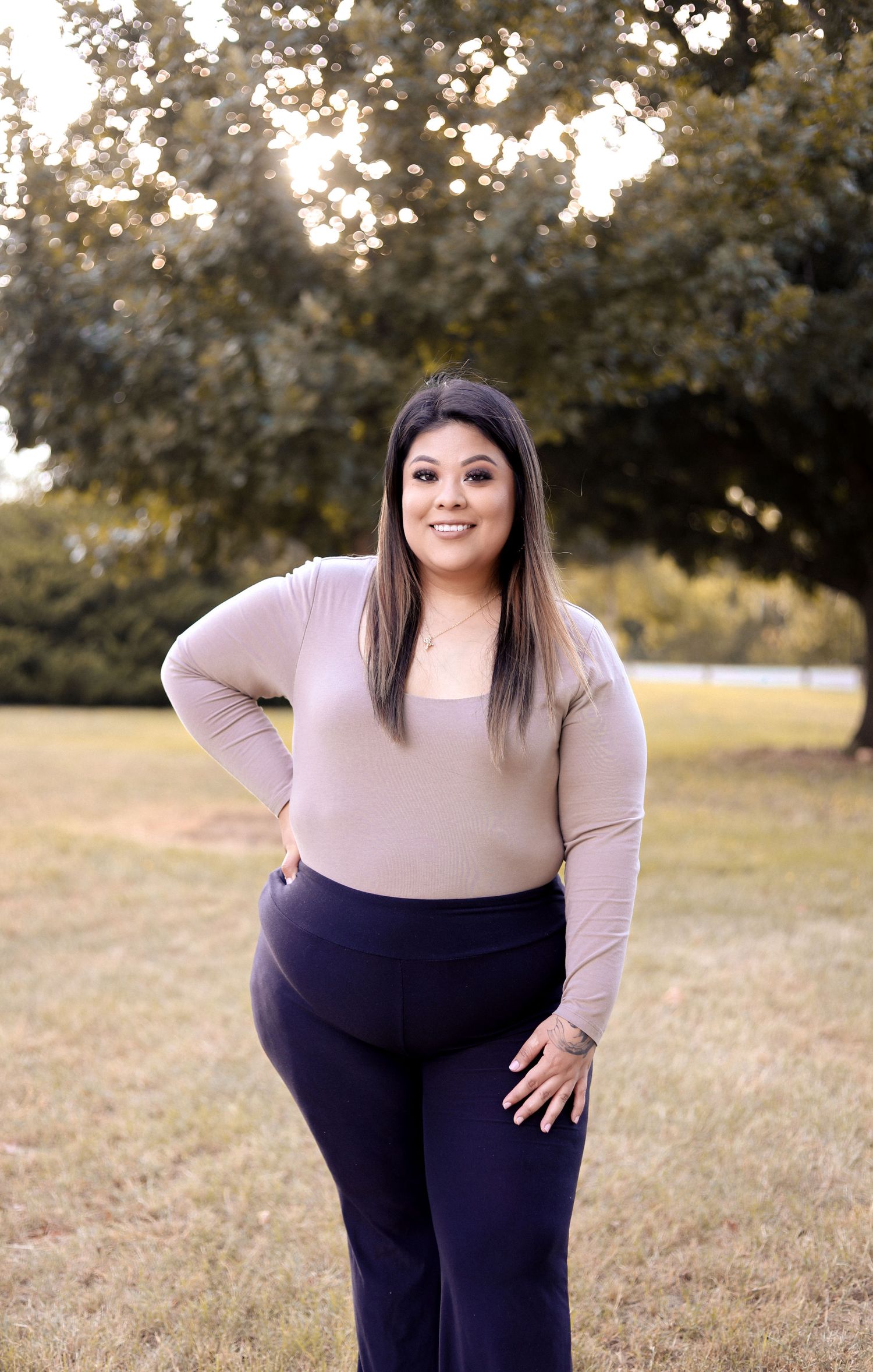 A smiling woman with long dark hair poses outdoors in a taupe top and navy pants with trees behind her.