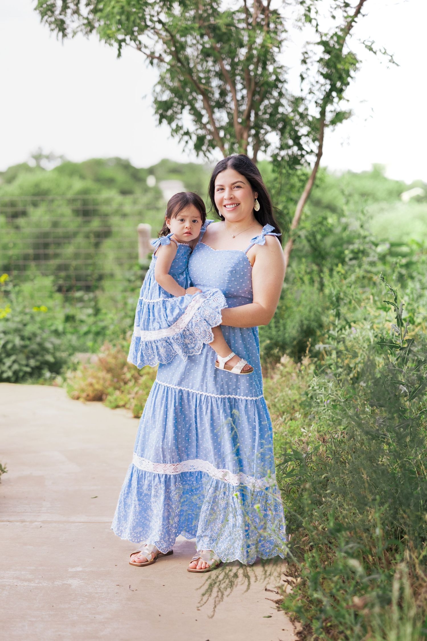 Mother and toddler daughter wear matching blue floral dresses while posing on a garden pathway.