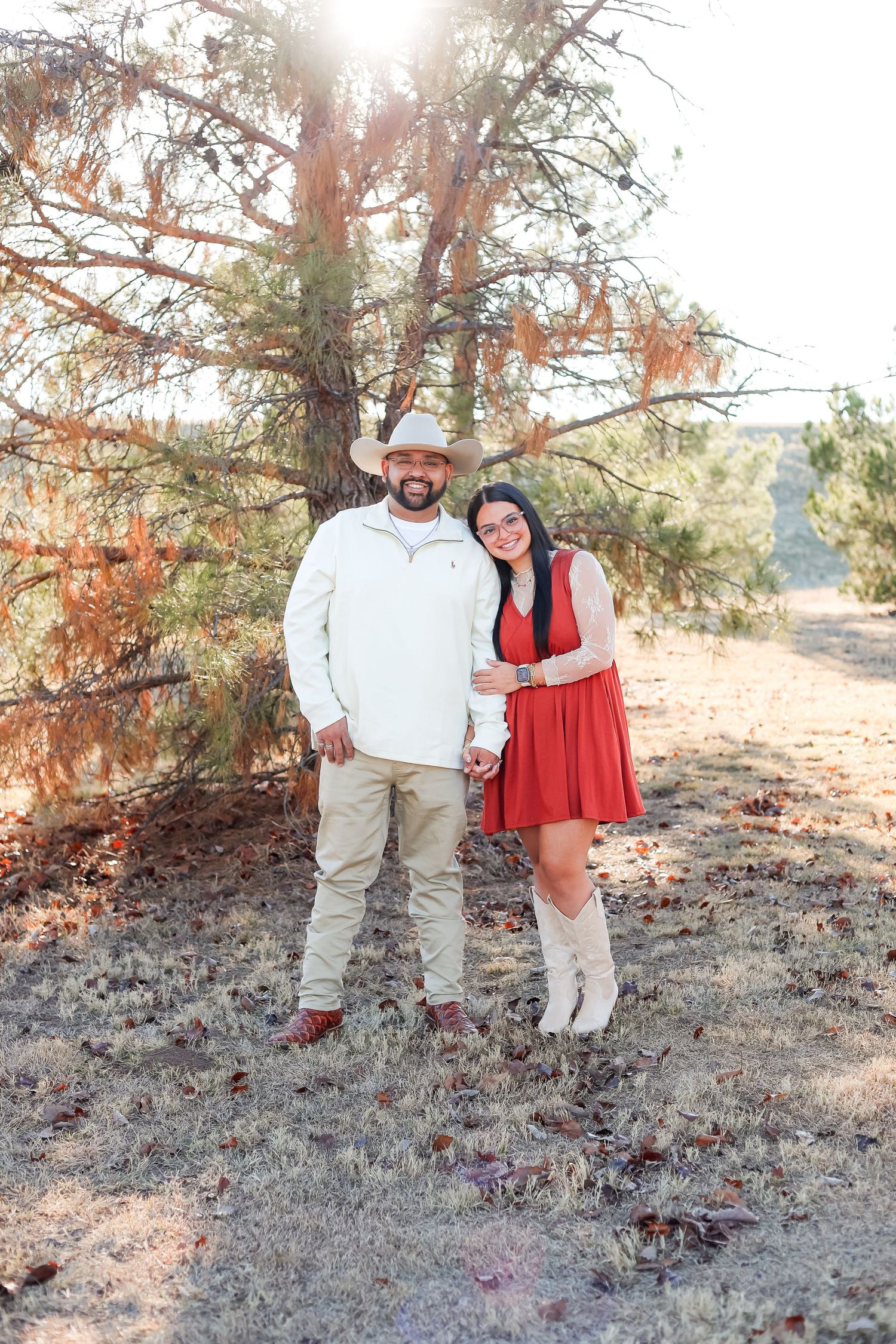 Couple posing outdoors under a pine tree, man in cowboy hat and white sweater, woman in red dress and boots.