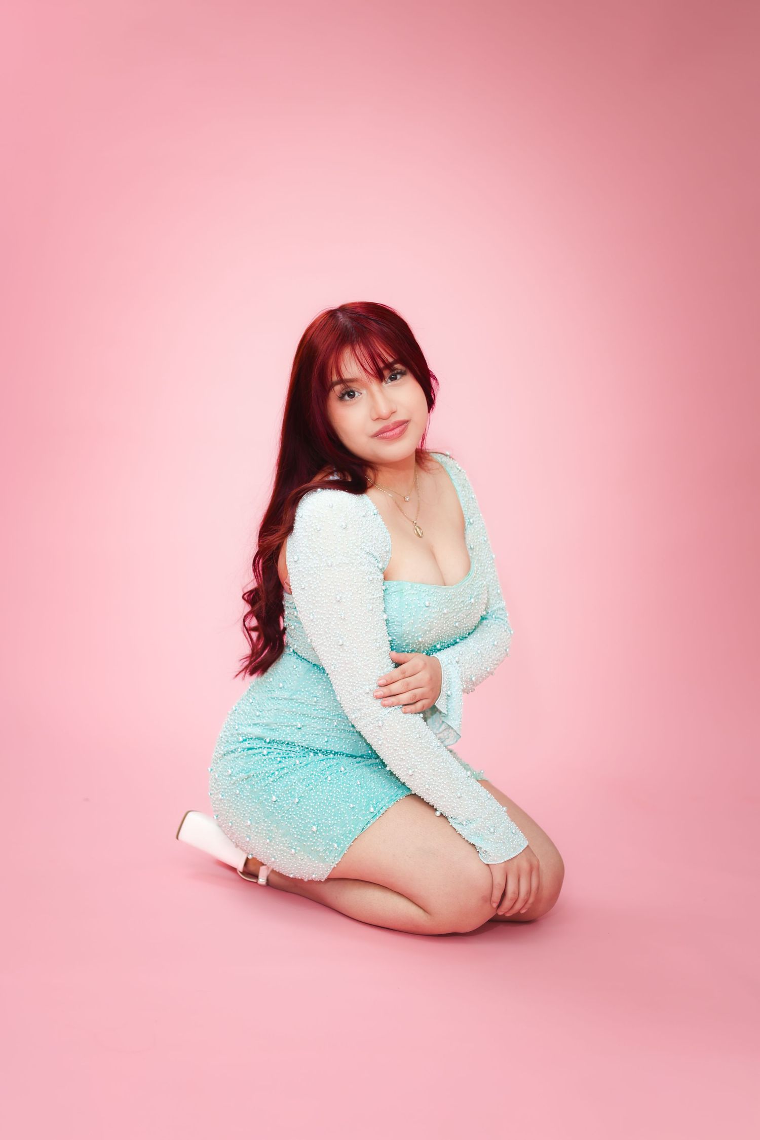 A woman in a teal sparkly dress poses gracefully on her knees against a soft pink studio background.