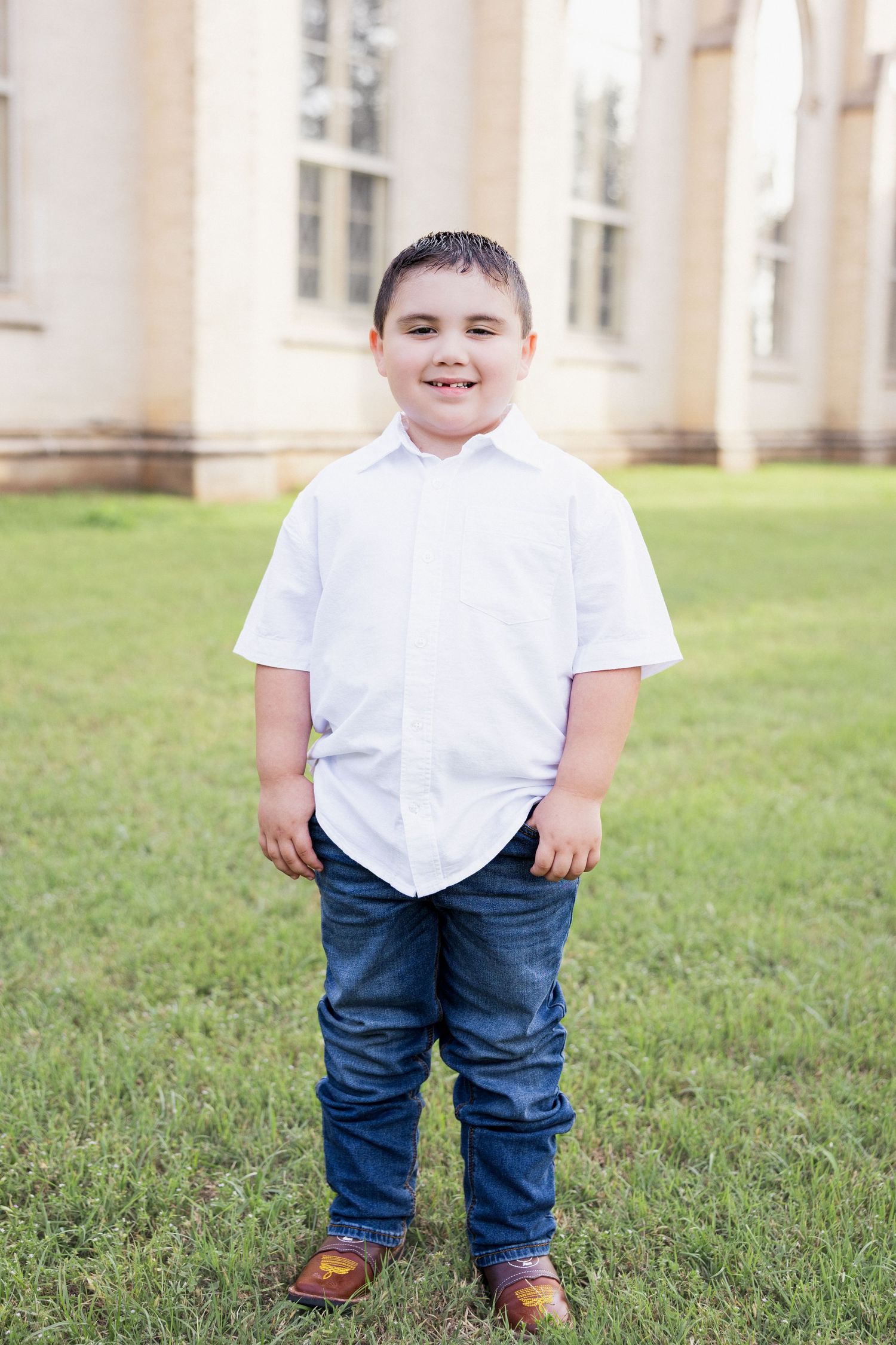 Smiling young boy in white button-up shirt and jeans poses on green grass near a building.