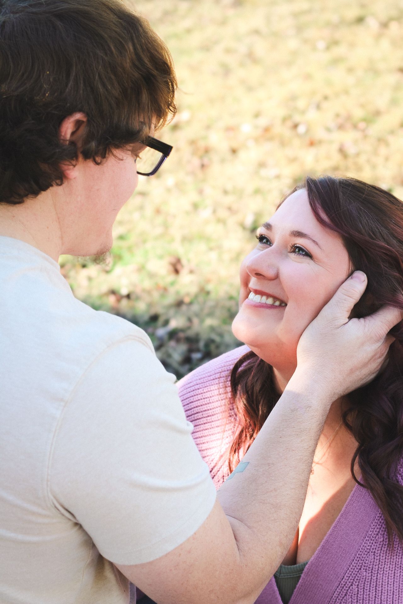 A smiling woman gazes up at a man with glasses who gently cups her face outdoors in a romantic moment.