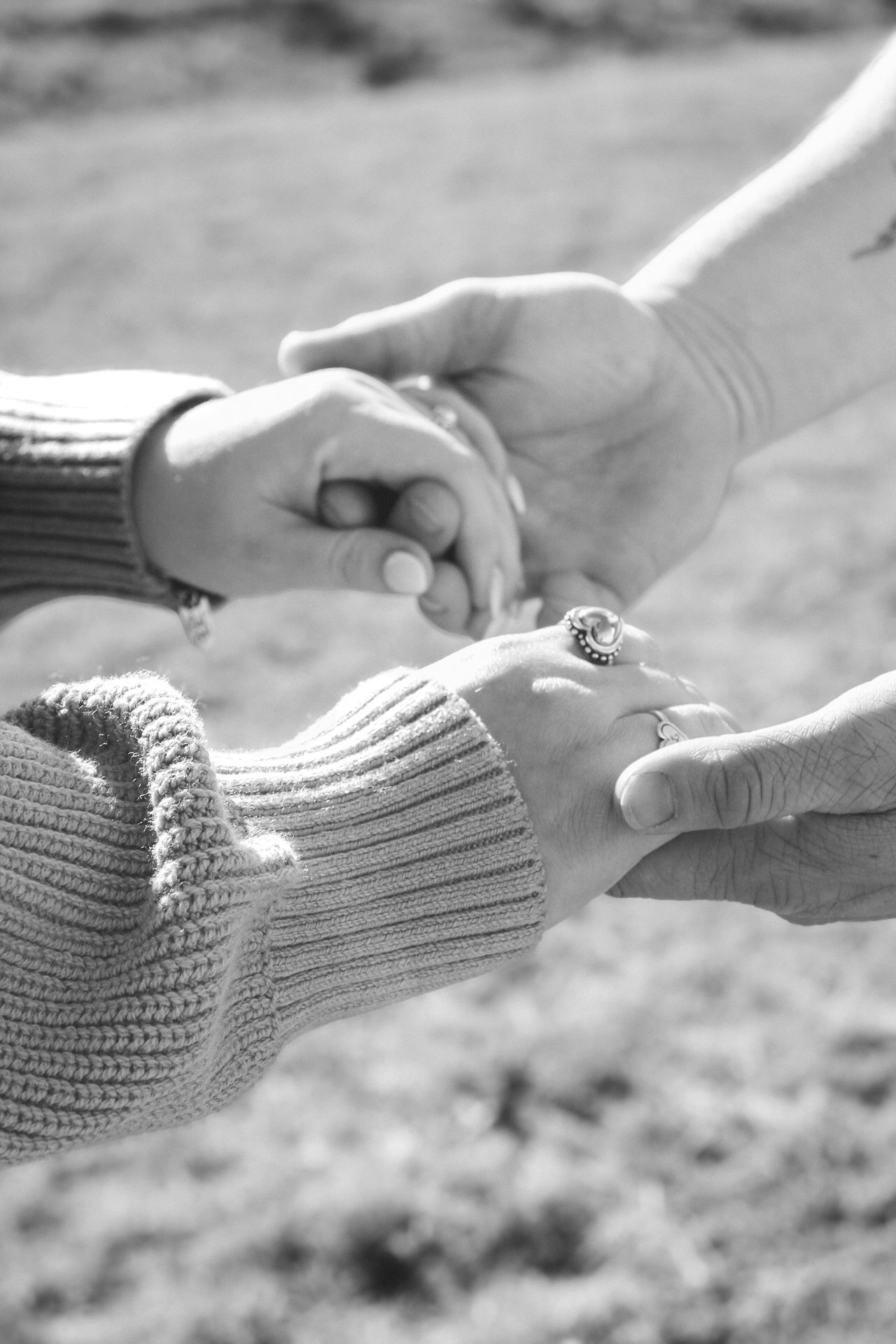 Black and white close-up of two people holding hands outdoors, wearing rings and knitwear.