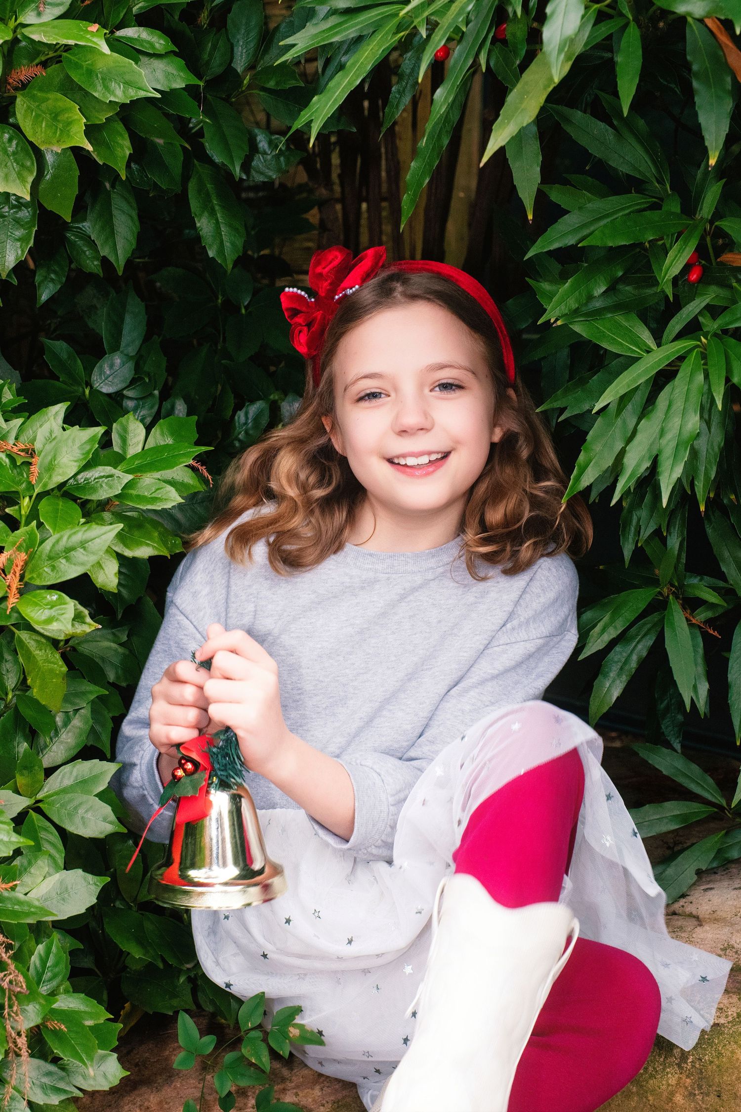 Child in gray sweater holds Christmas ornaments and bells against green foliage backdrop.