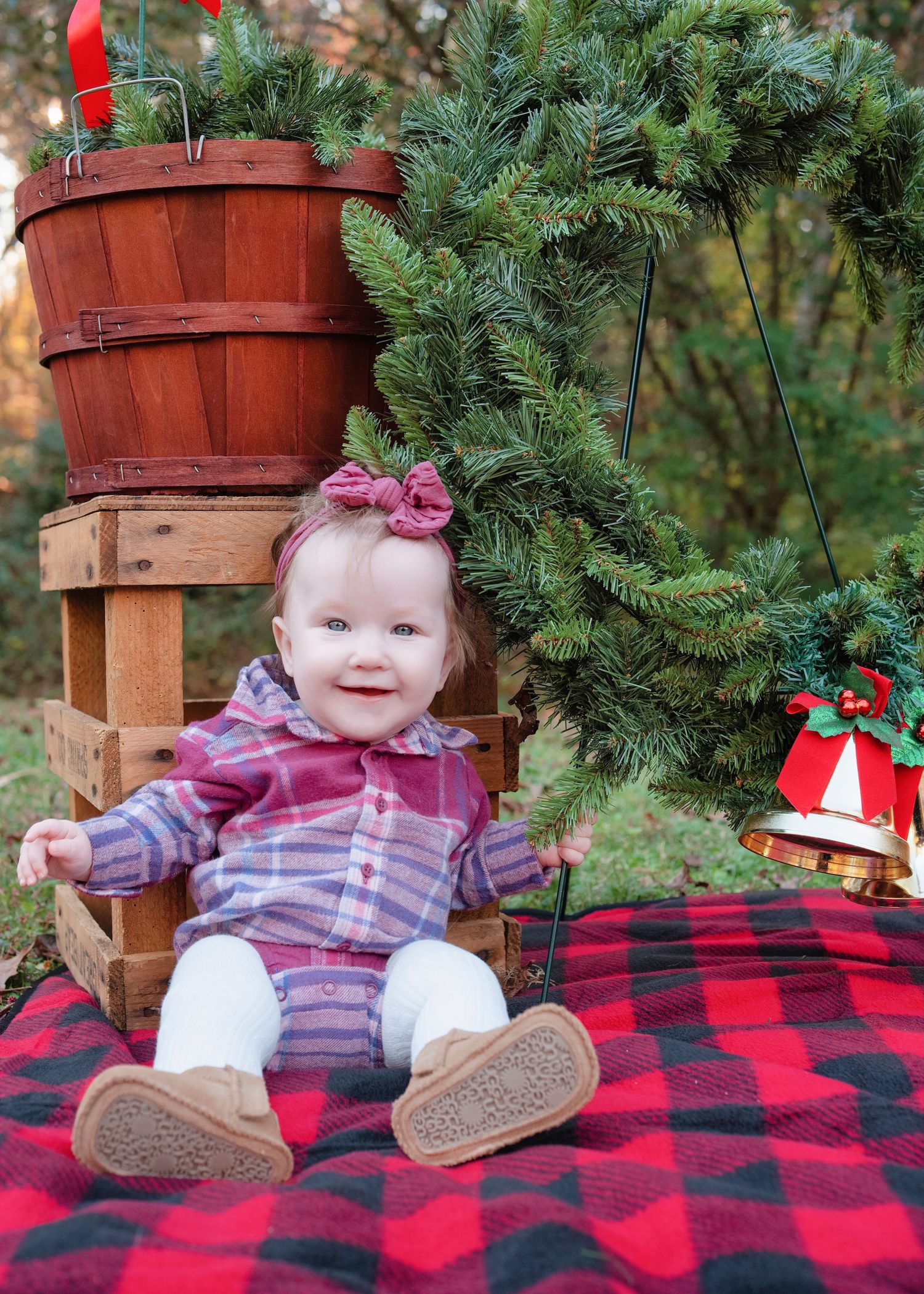 Baby in plaid outfit sits on red buffalo check blanket with Christmas decorations and greenery.
