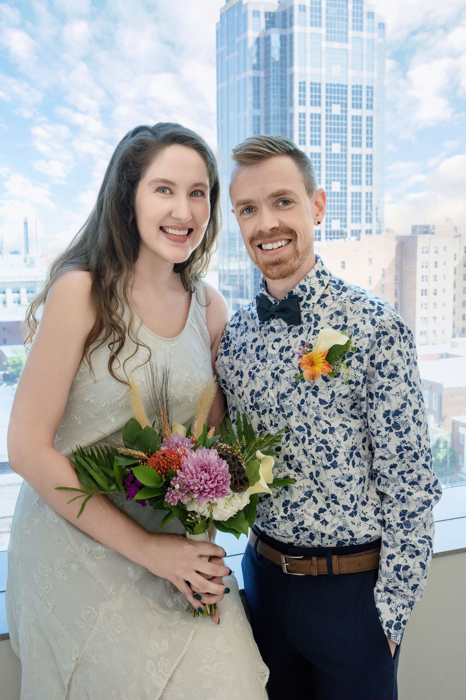 A couple smiles together at a city venue with a skyscraper backdrop and floral bouquet.