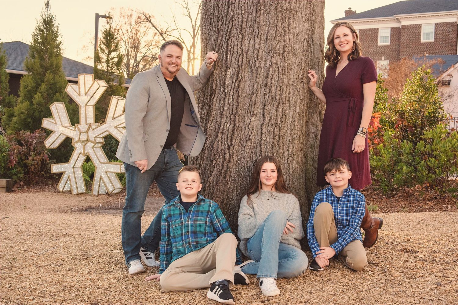 Family group photo sitting by large tree trunk with decorative snowflake prop.