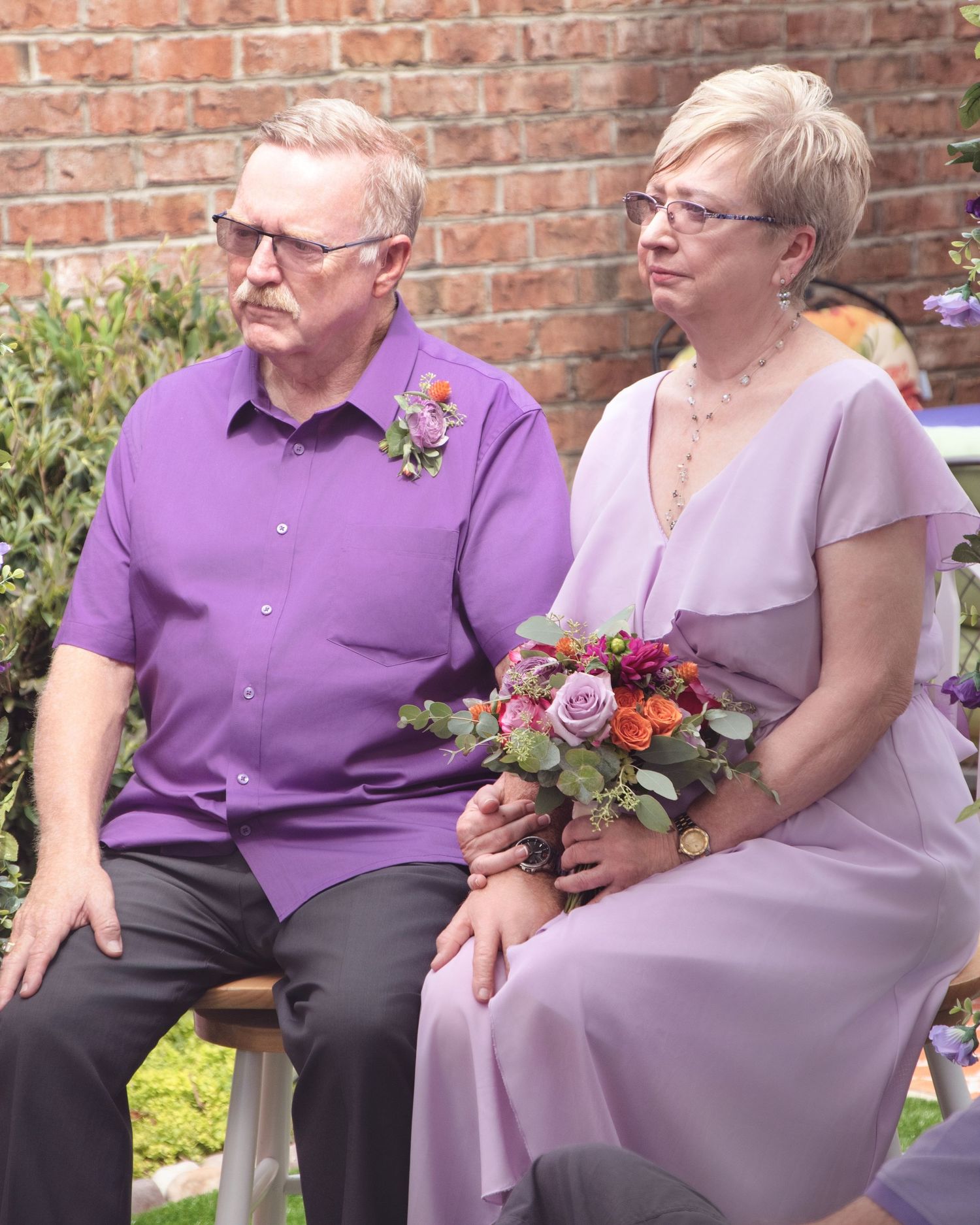 Couple in coordinating purple and lavender formal attire sits together on bench with floral bouquet against brick wall.