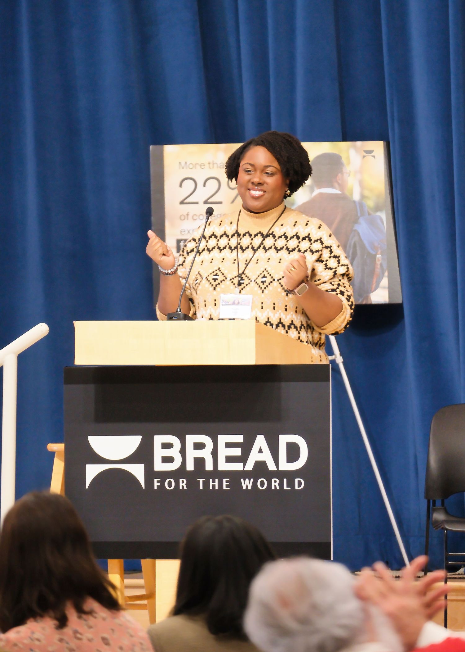 A speaker wearing a patterned sweater gestures enthusiastically at a podium with the Bread for the World logo.