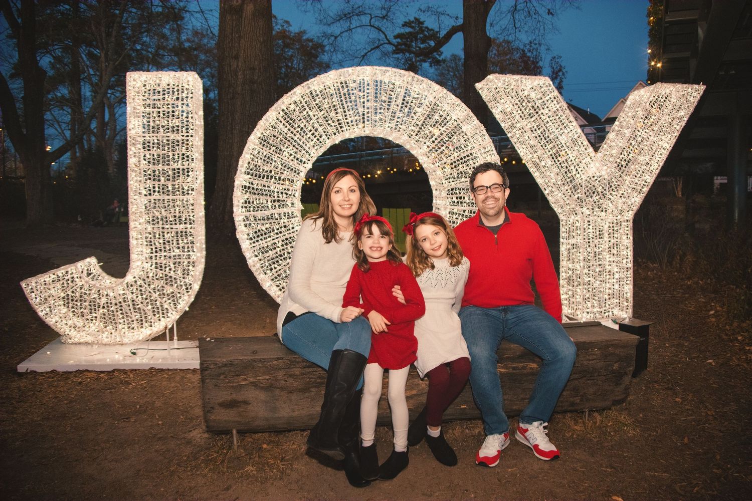 A family poses together in front of a large illuminated JOY holiday sign.