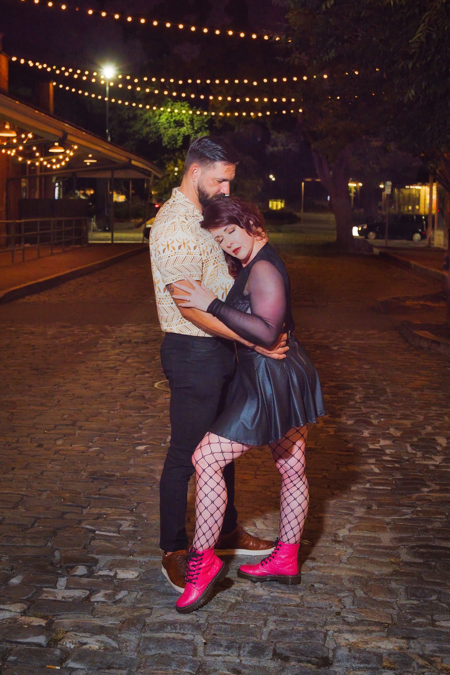 A couple embraces under string lights on cobblestone street at night.