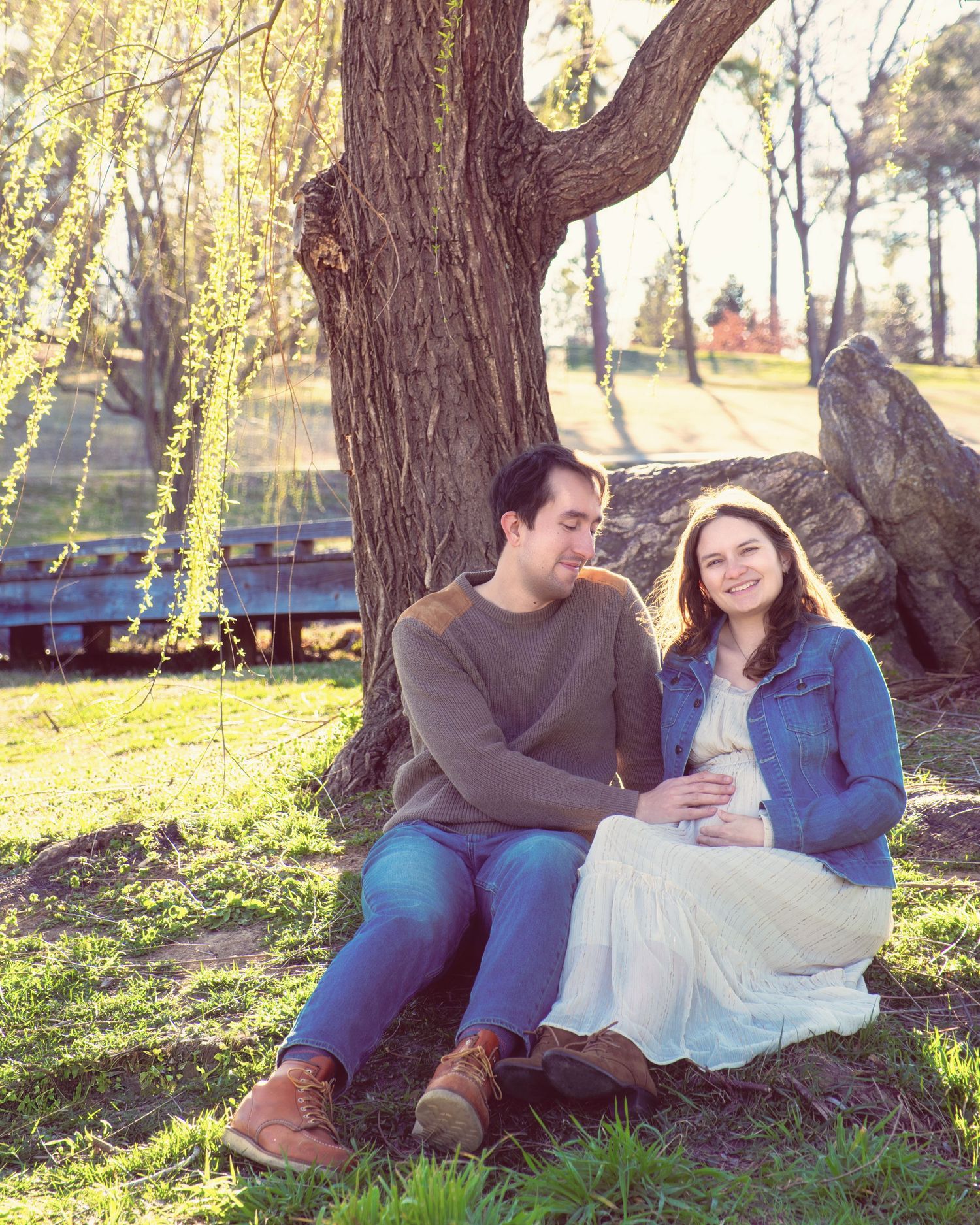 A couple sits together by a tree trunk in a sunlit park on an autumn afternoon.