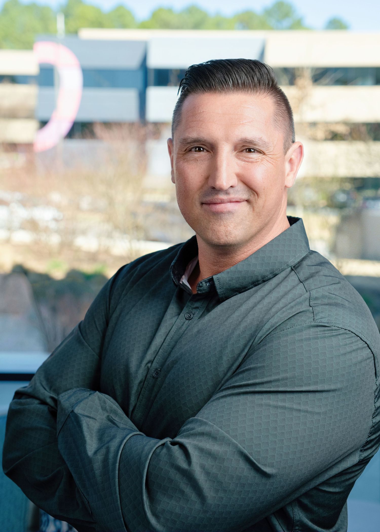 Professional headshot of person in dark colored polo shirt with arms crossed against blurred background outdoors.