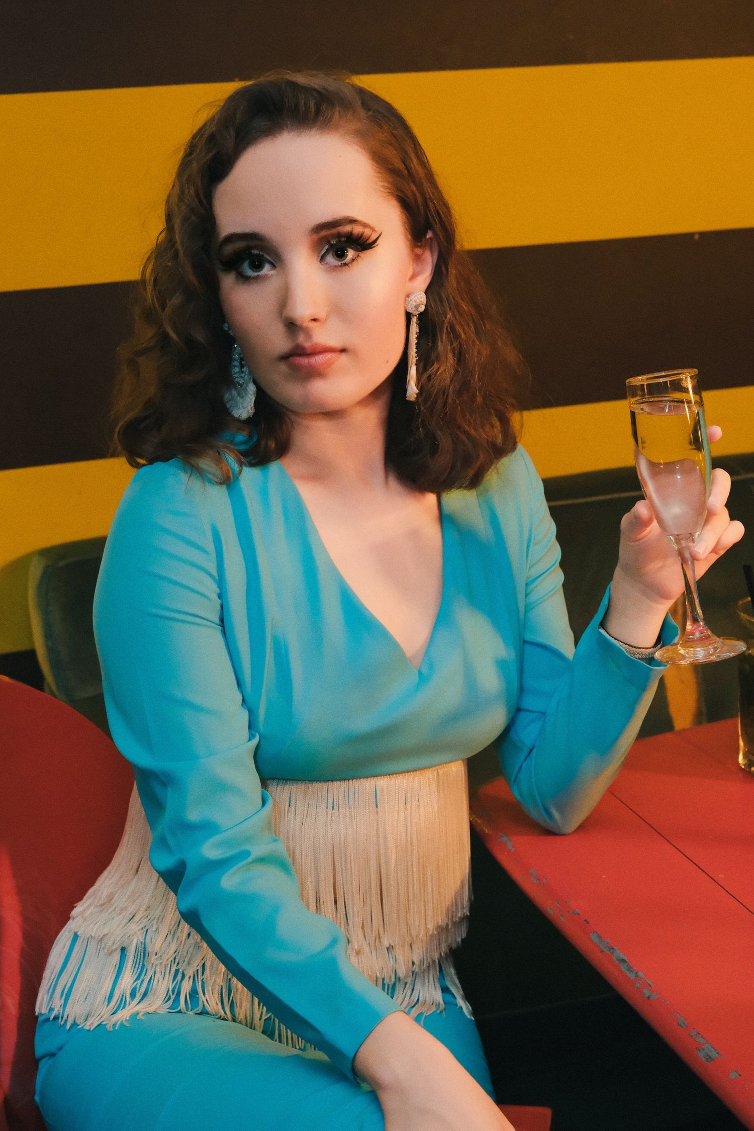 Glamorous portrait in a turquoise dress holding champagne against striped yellow and black background.